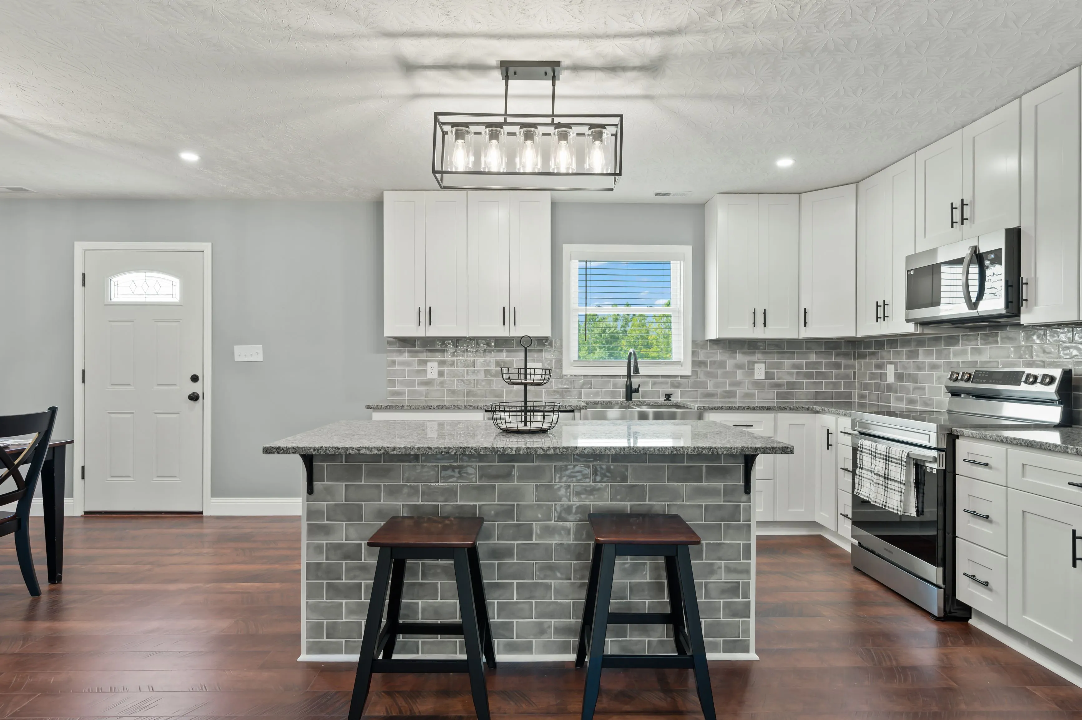 Modern kitchen interior with white cabinetry, stainless steel appliances, subway tile backsplash, granite countertops, and a kitchen island with two stools.