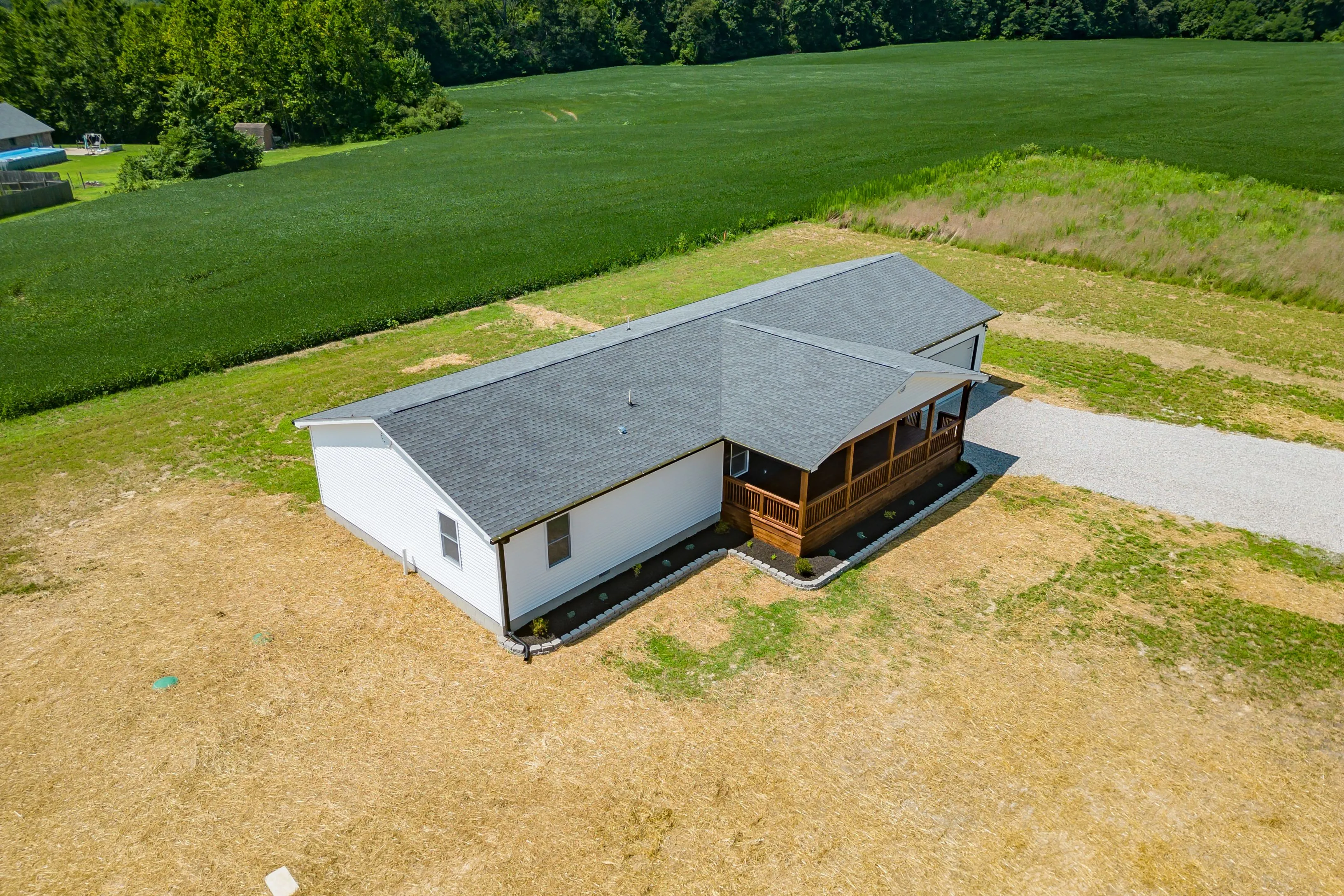 Aerial view of a modern single-story house with white siding and a dark roof, featuring a wooden porch, surrounded by a large grassy field and a gravel driveway.