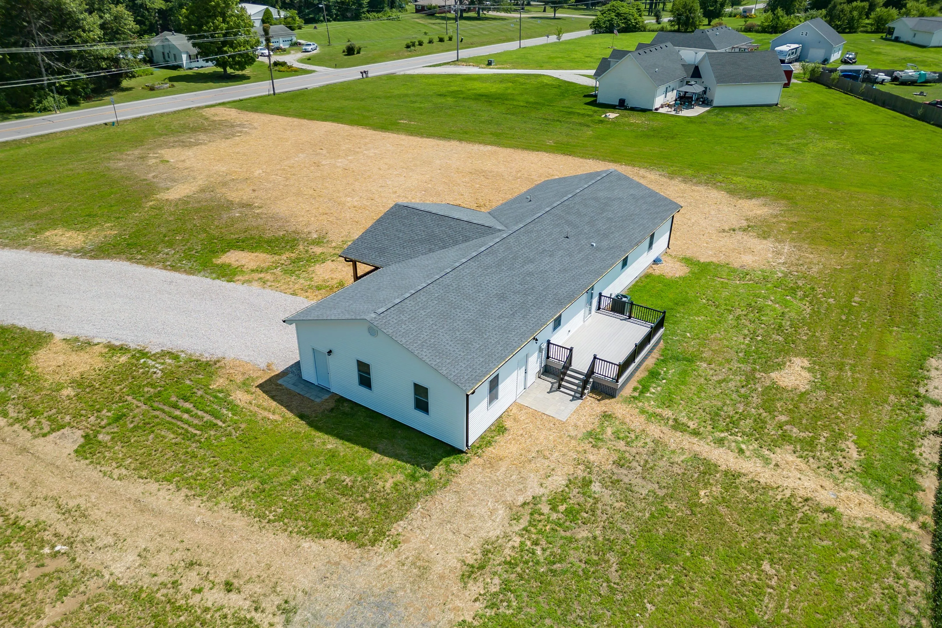 Aerial view of a long, single-story building with a gray shingled roof and white siding, featuring a small deck with patio furniture, surrounded by a patchy grass field and a gravel driveway, with other residential properties in the background.