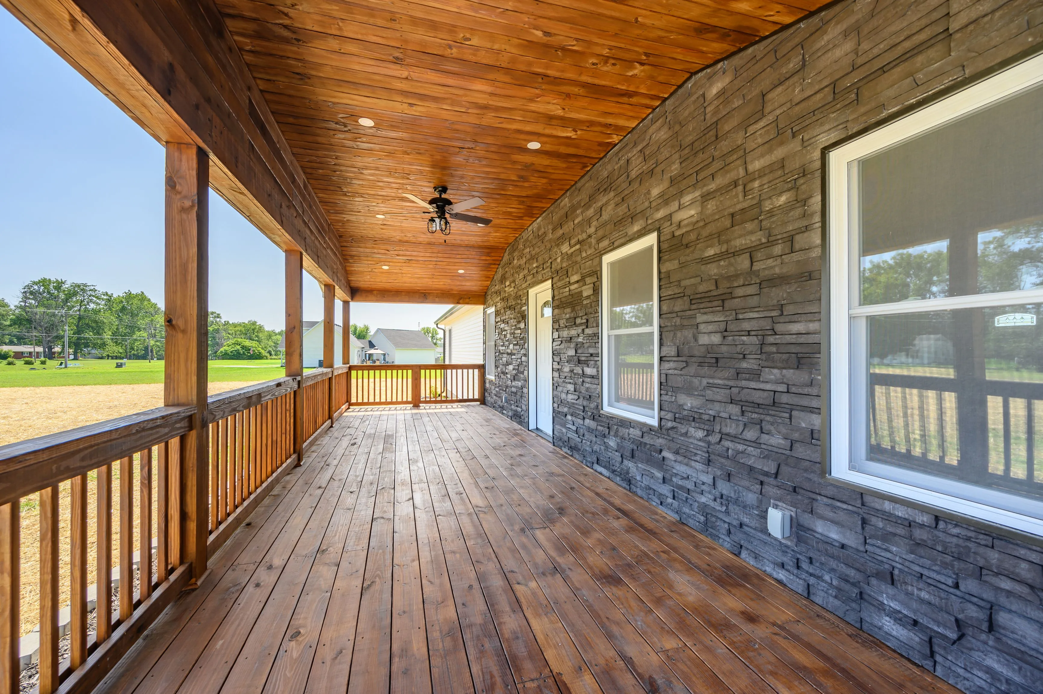 Spacious wooden porch with a natural wood ceiling, stone accent wall, ceiling fan, and a view of a sunny backyard.