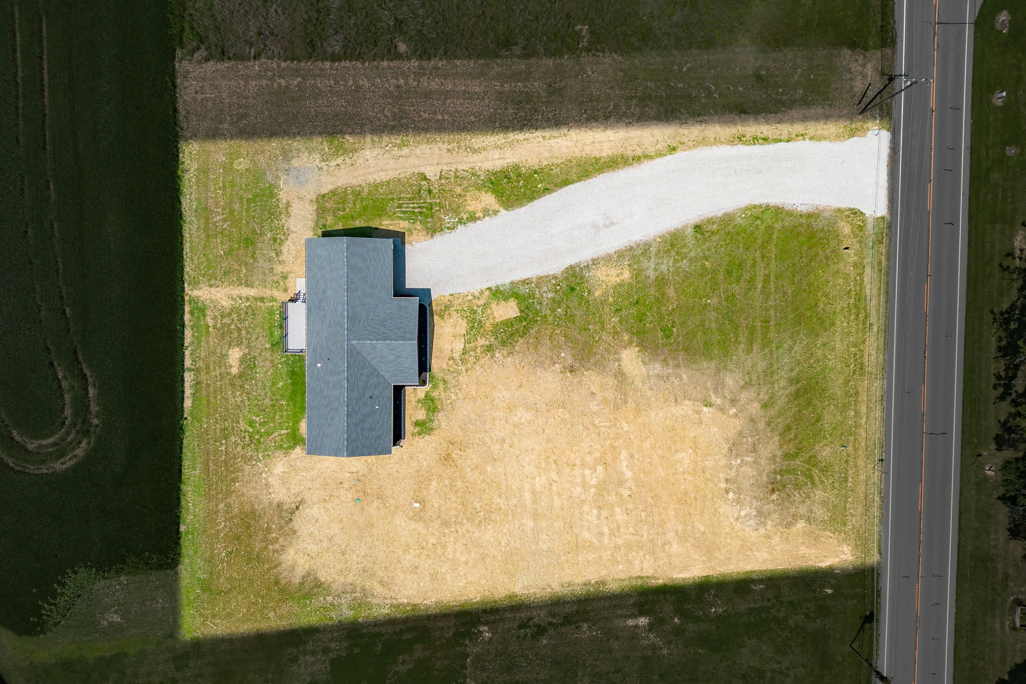 Aerial view of a house with a gray roof next to a country road, surrounded by patches of green grass and fields.
