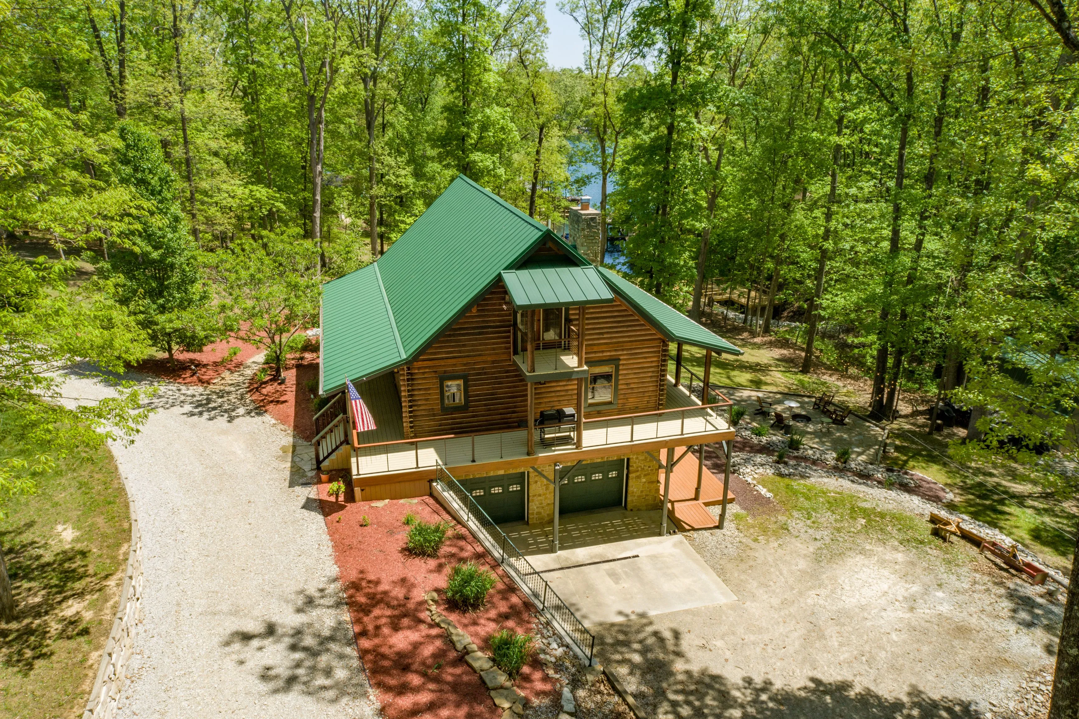 Aerial view of a secluded cabin with a green roof surrounded by lush green trees with a gravel driveway.
