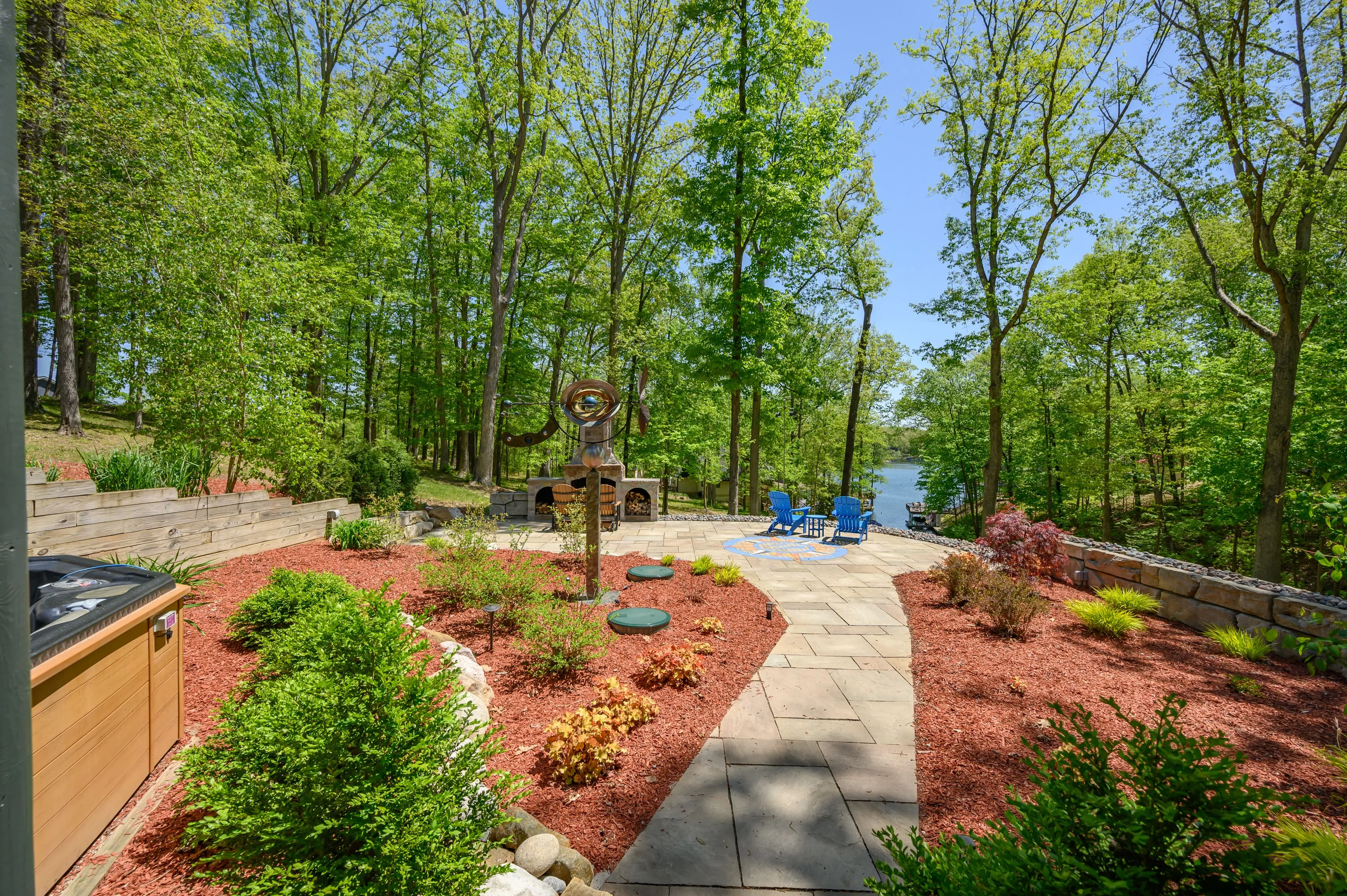 Beautifully landscaped backyard with a stone pathway, hot tub, outdoor fireplace, and two Adirondack chairs overlooking a tranquil lake surrounded by lush greenery.