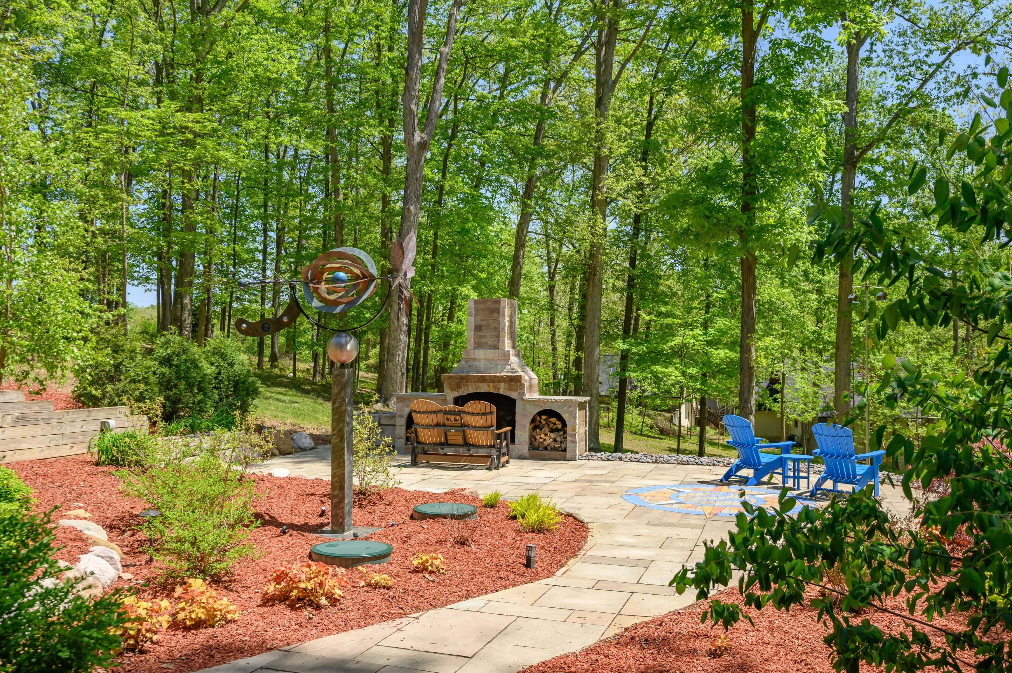 A serene backyard with lush trees, a stone footpath, a sculptural wind spinner, an outdoor fireplace, and two blue Adirondack chairs.