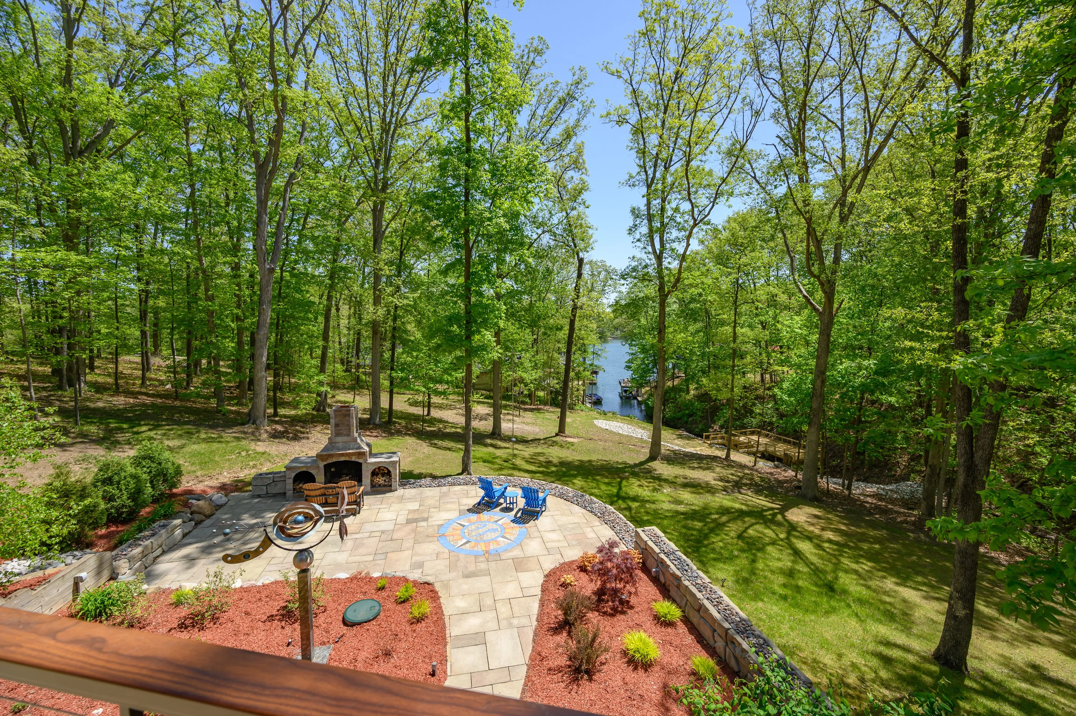 Backyard patio with a fire pit and seating area surrounded by lush green forest and a view of a lake in the distance.