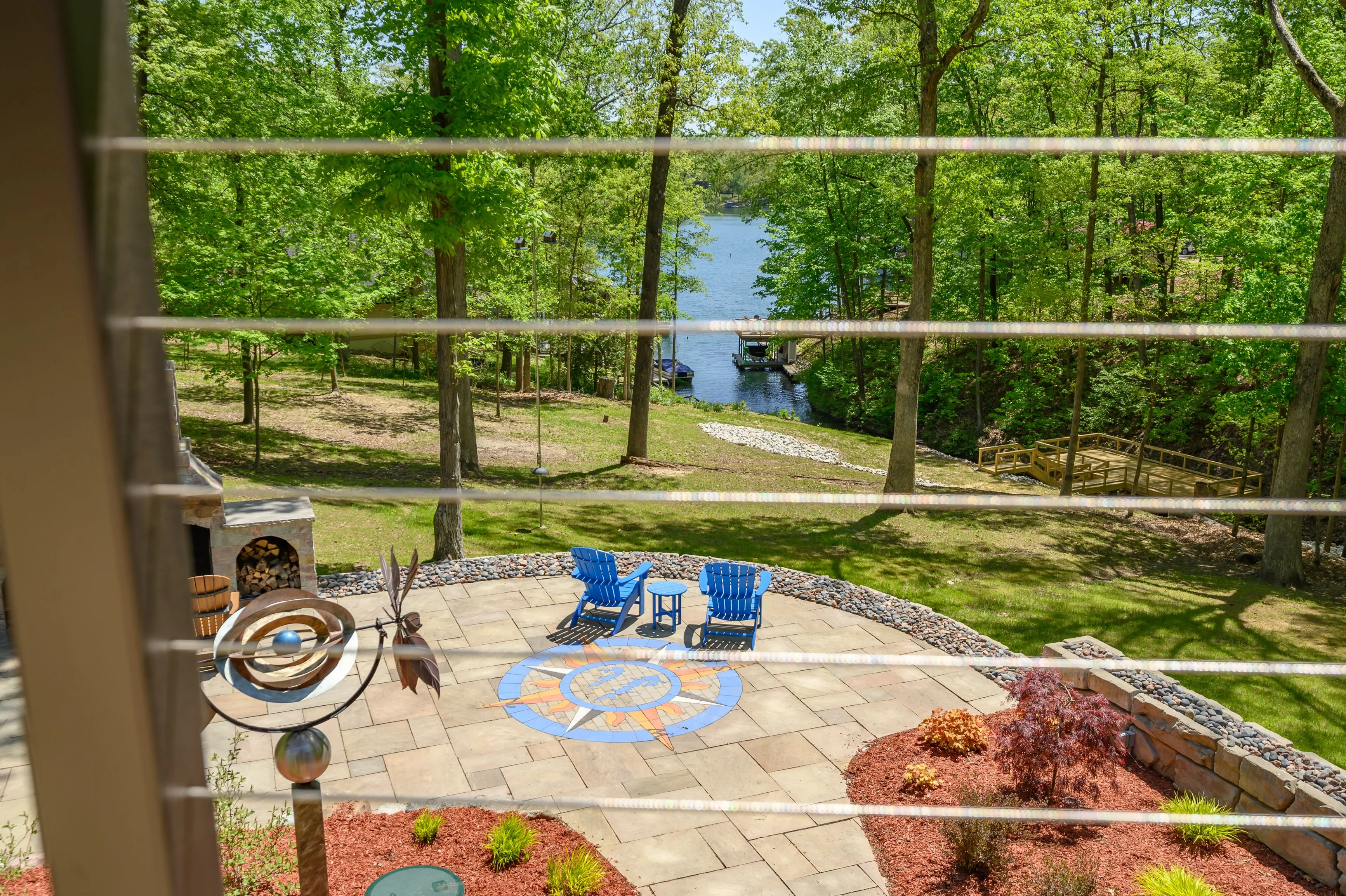 View of a patio with chairs and a fire pit through a window, overlooking a garden and lake in the distance.