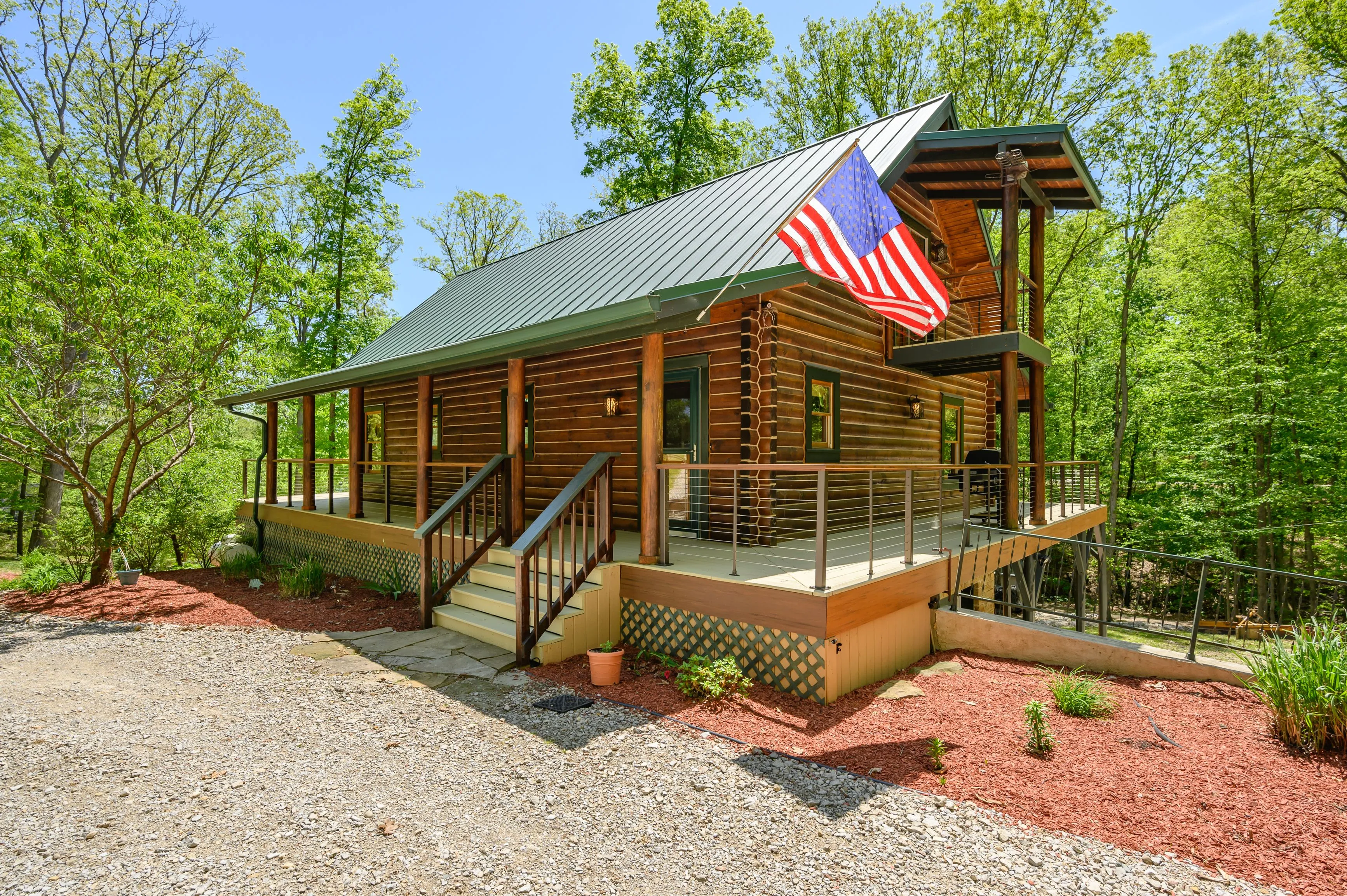 Wooden log cabin with an American flag flying, surrounded by trees under a clear blue sky.