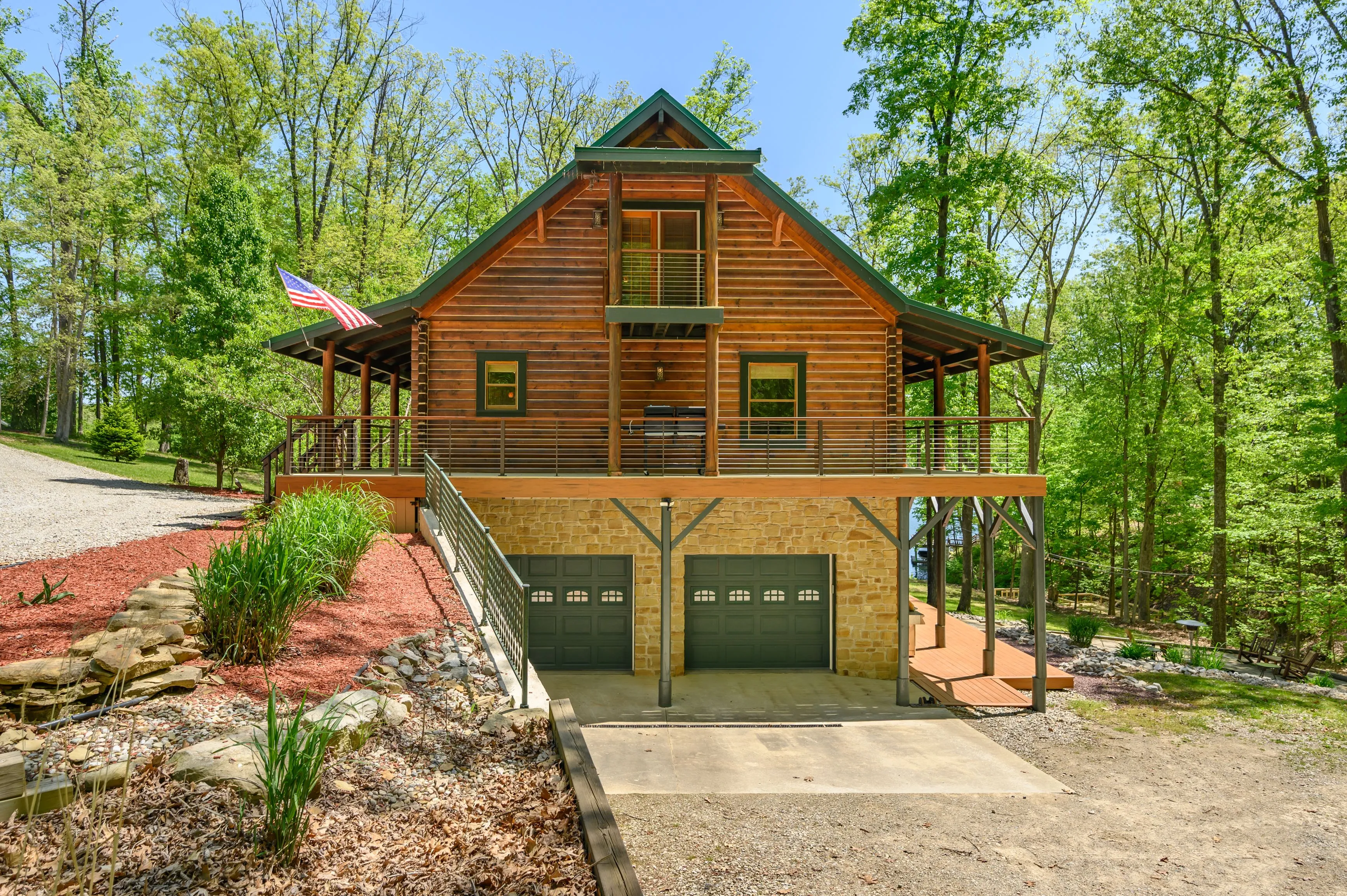A wooden cabin with a spacious balcony, surrounded by green trees, featuring a stone foundation and a three-car garage beneath, with an American flag on display.