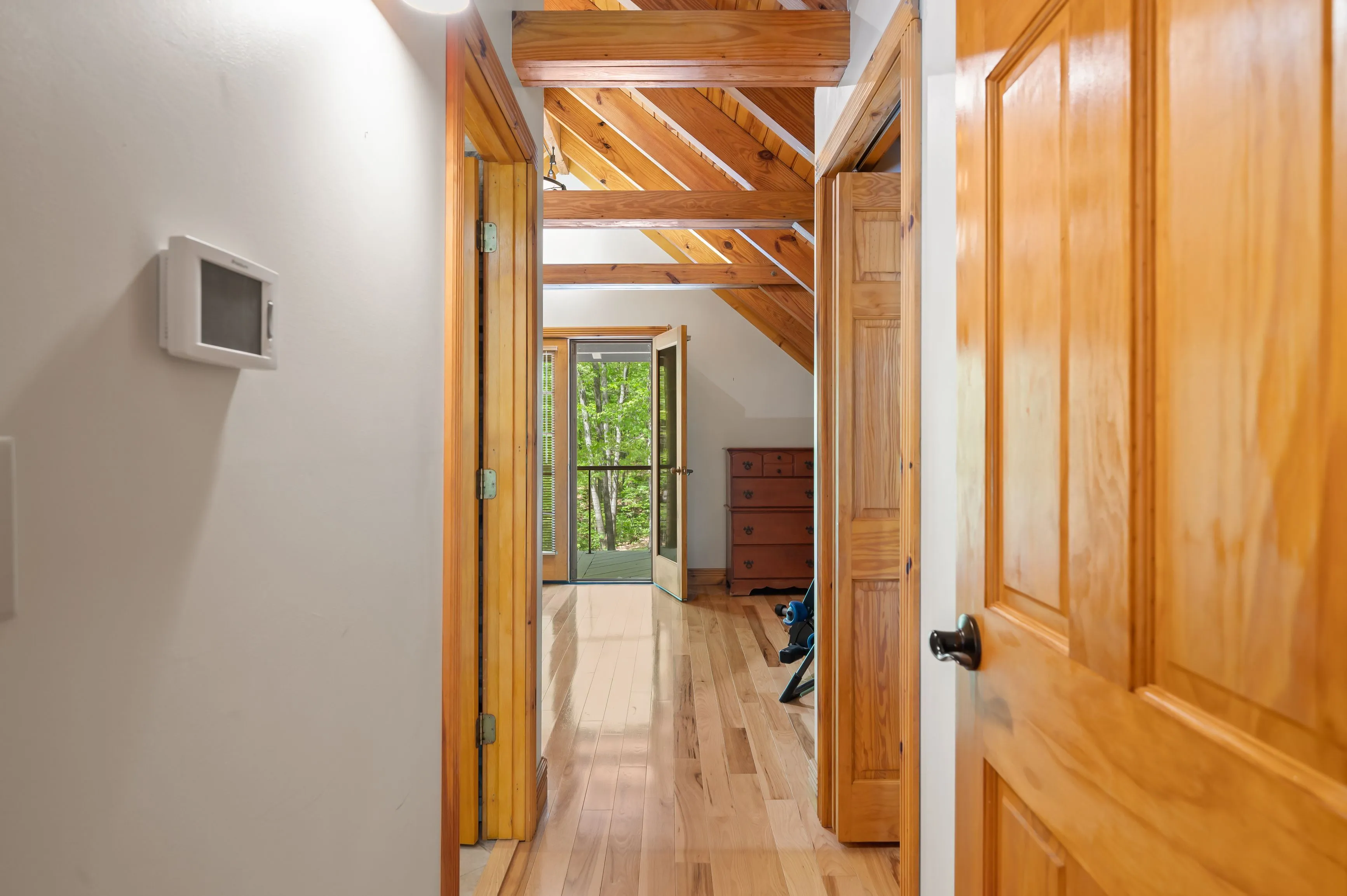 Interior view of a hallway with hardwood floors, wooden doors, and exposed wooden beams, leading towards a room with natural light.