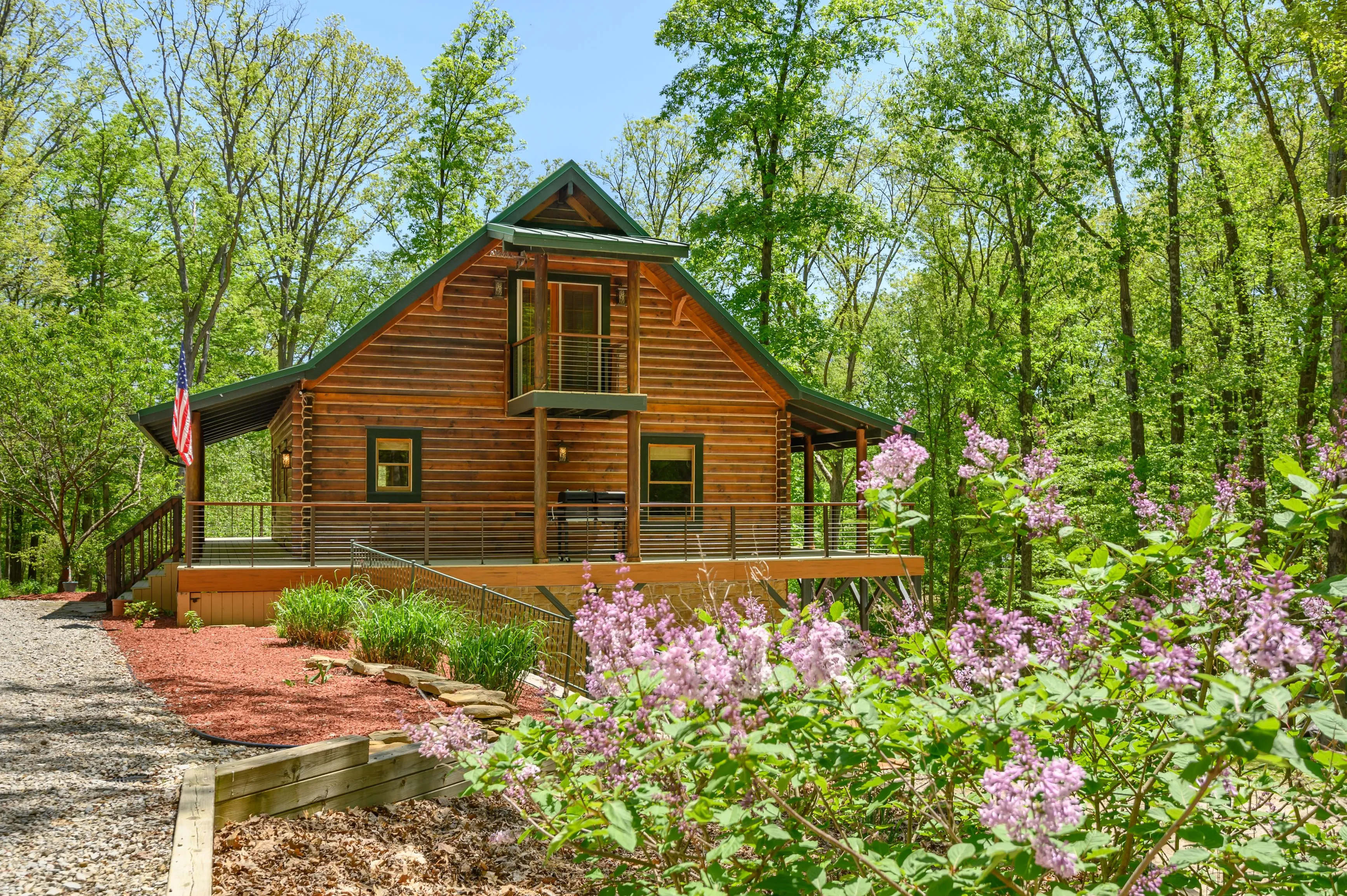Wooden cabin surrounded by forest with blooming lilac flowers in the foreground.