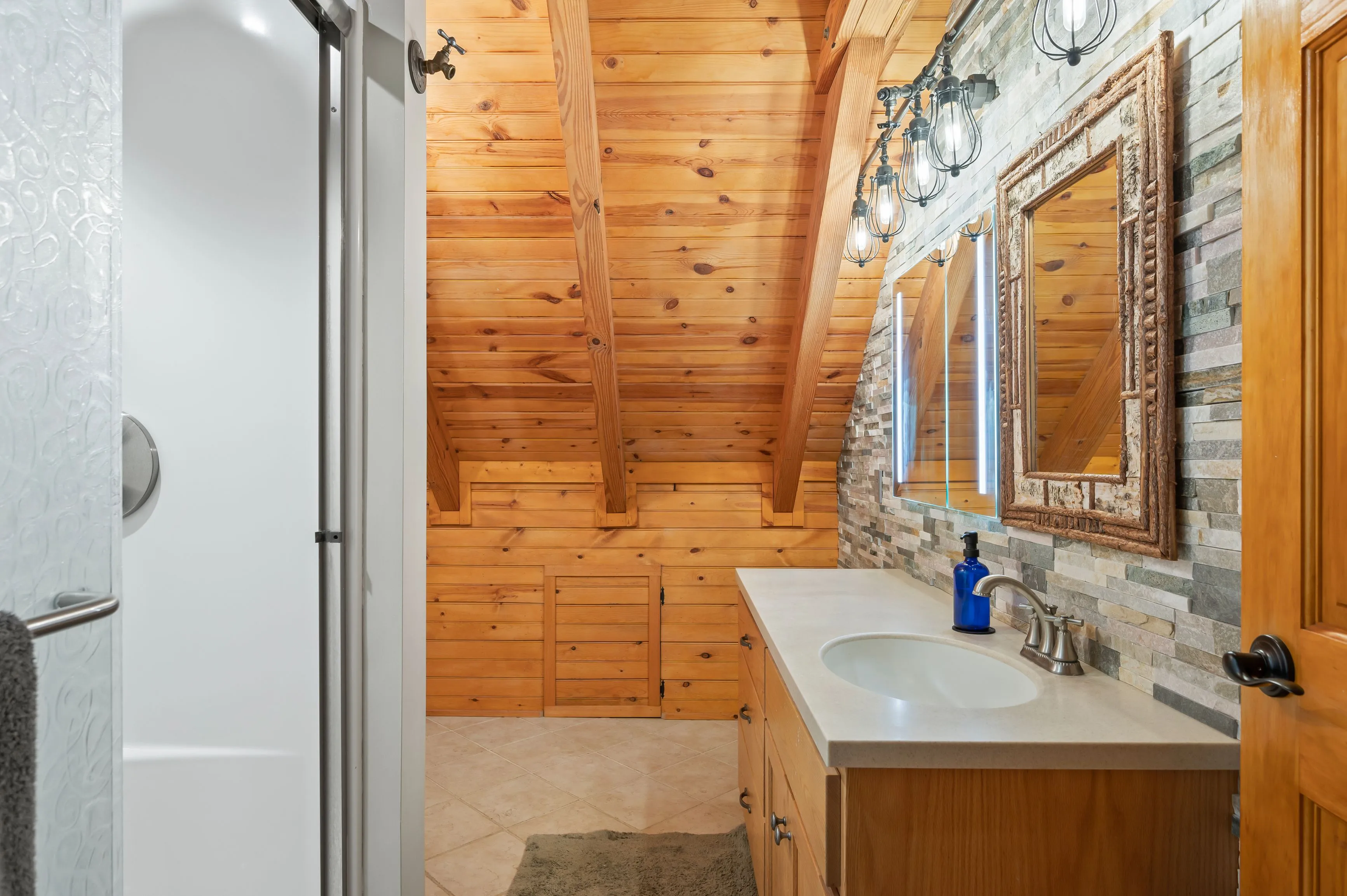 Interior of a rustic bathroom with wooden walls, a white sink, mirror, and a glass shower door.