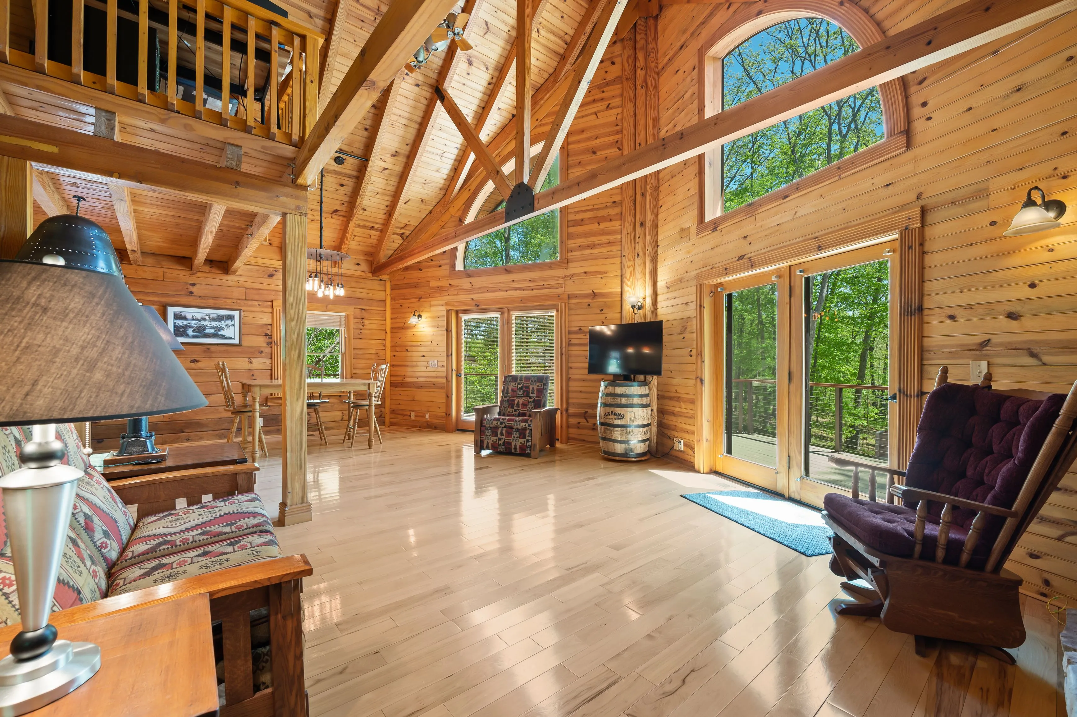 Cozy wooden cabin interior with high ceilings, large windows, and rustic furniture, featuring a loft area and polished wood floors.