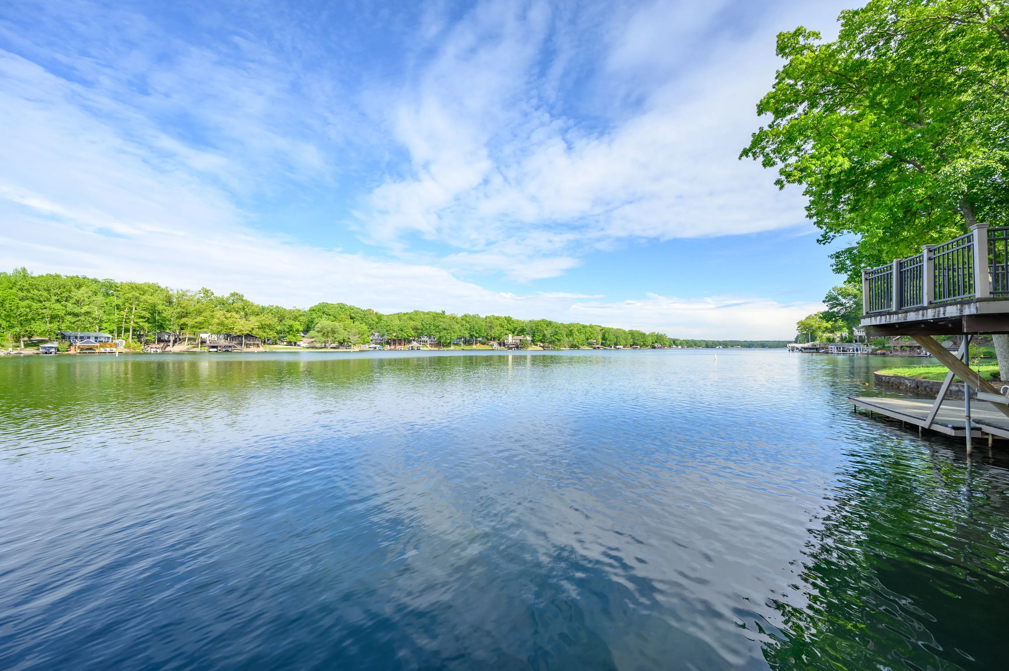 Tranquil lake surrounded by greenery under a vast blue sky with scattered clouds, a pier visible to the right.
