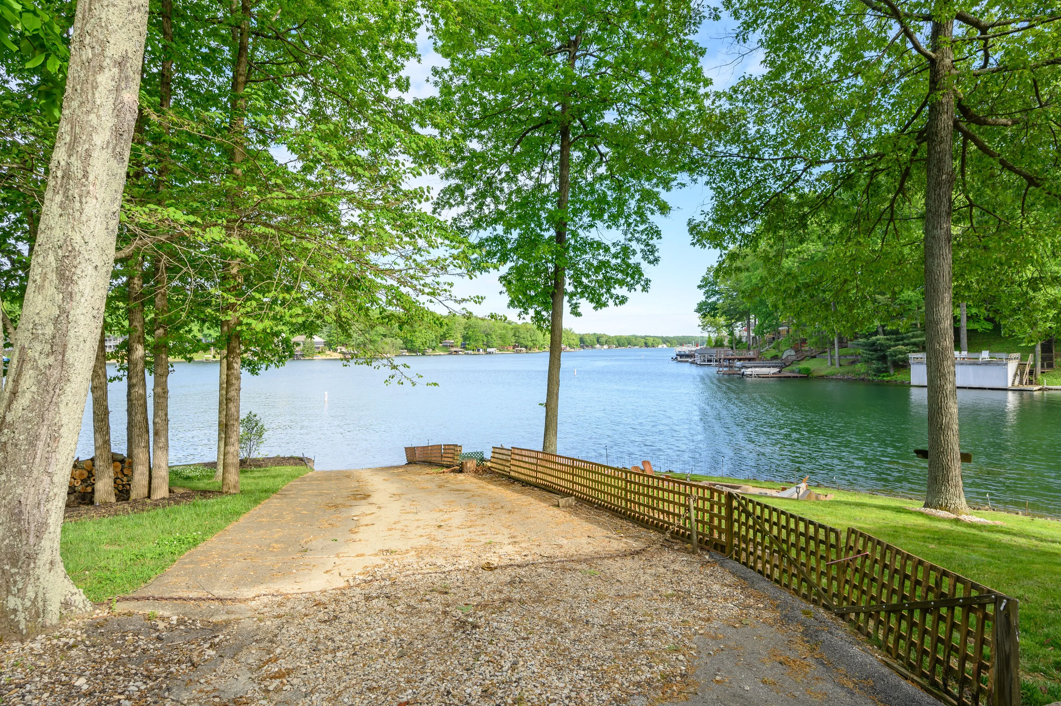 Gravel path leading to a tranquil lake surrounded by green trees with a wooden fence on one side.