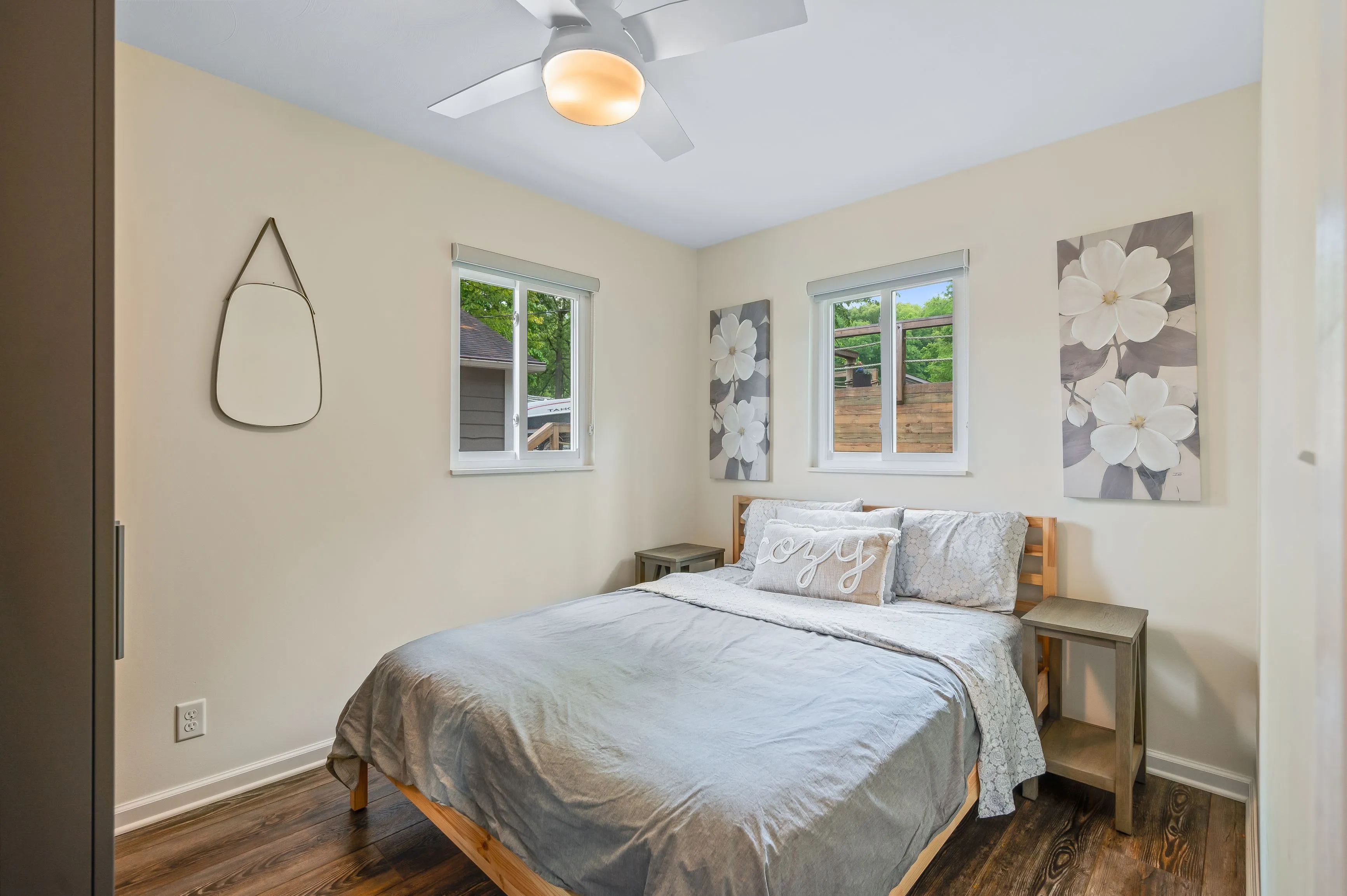 Bright cozy bedroom with a double bed, white linens, wooden bedside bench, light walls, two windows, and a ceiling fan.