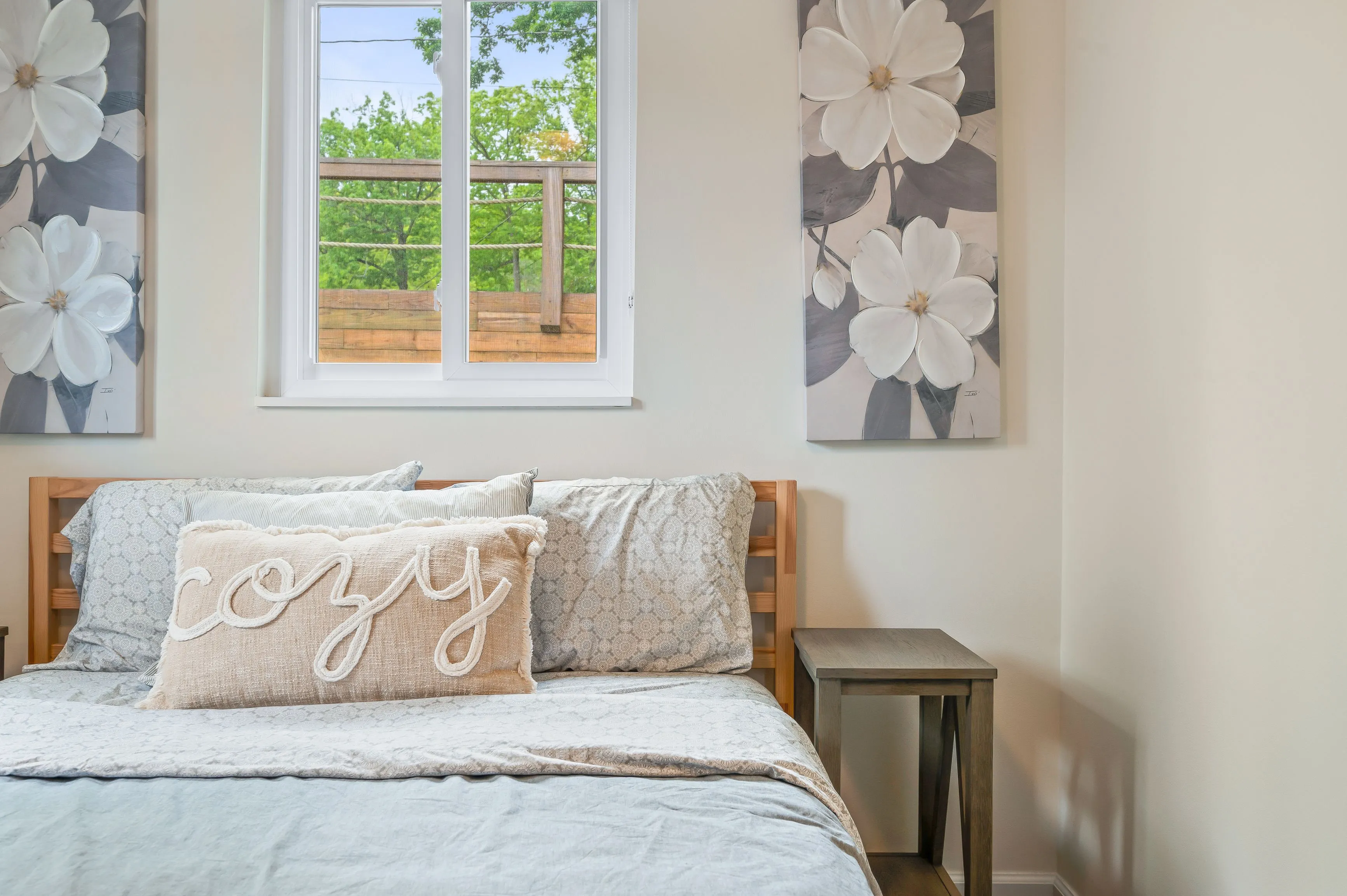 Cozy bedroom interior with a bed covered in soft linens, decorative pillows, and a floral artwork on the wall next to a window with a garden view.