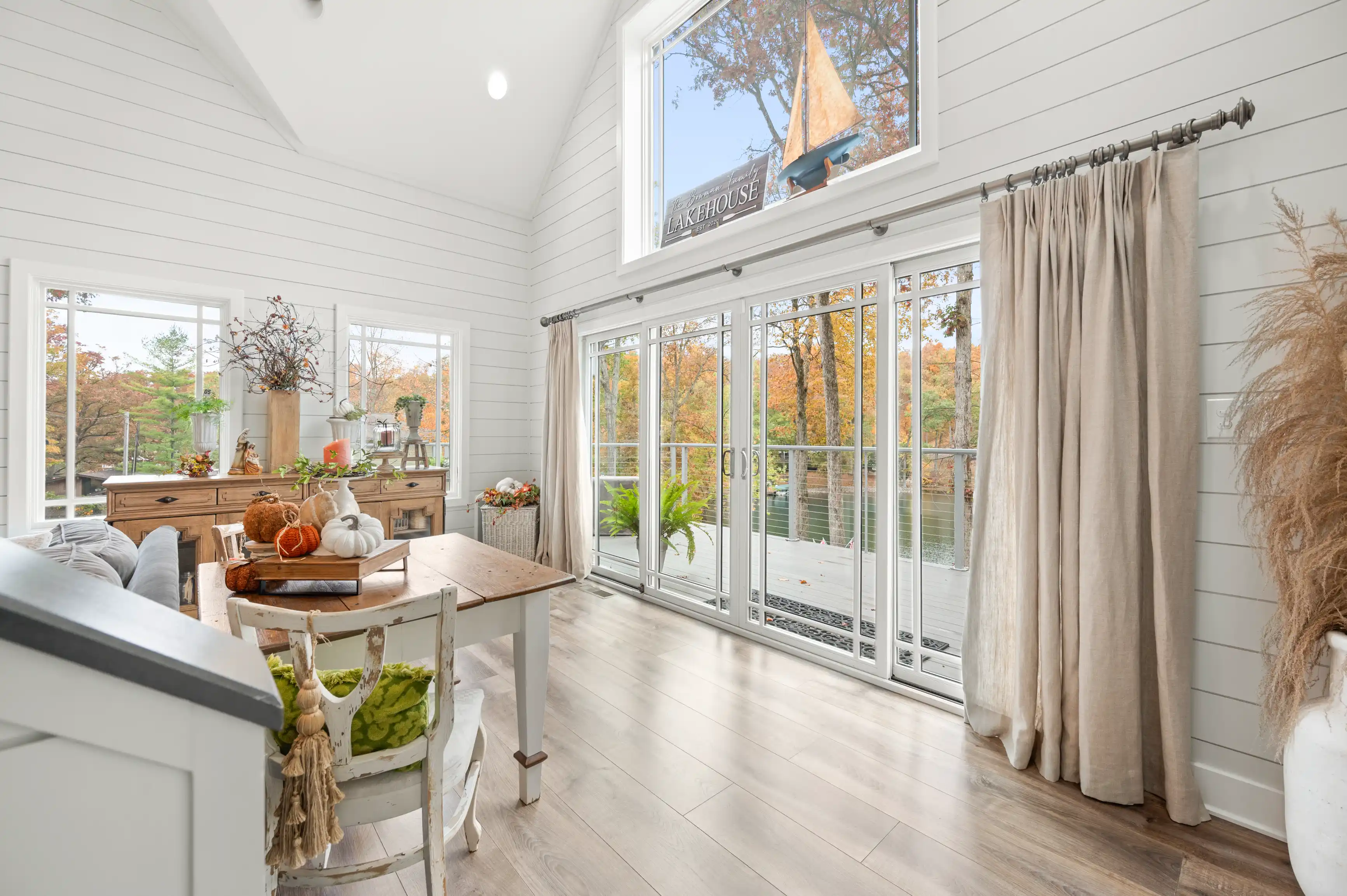 Bright and airy sunroom with shiplap walls and hardwood floors, tastefully decorated with autumn decor, featuring a view of trees with fall foliage through large glass doors.