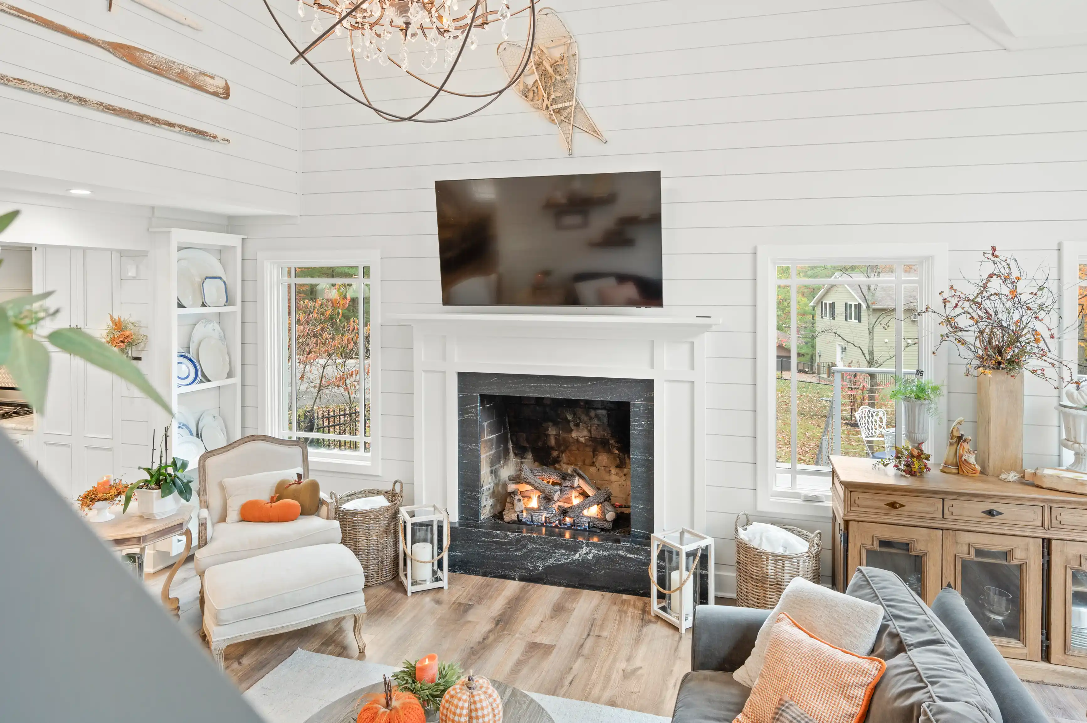 Cozy living room with white shiplap walls, a lit fireplace, autumn decorations, and a chandelier above.