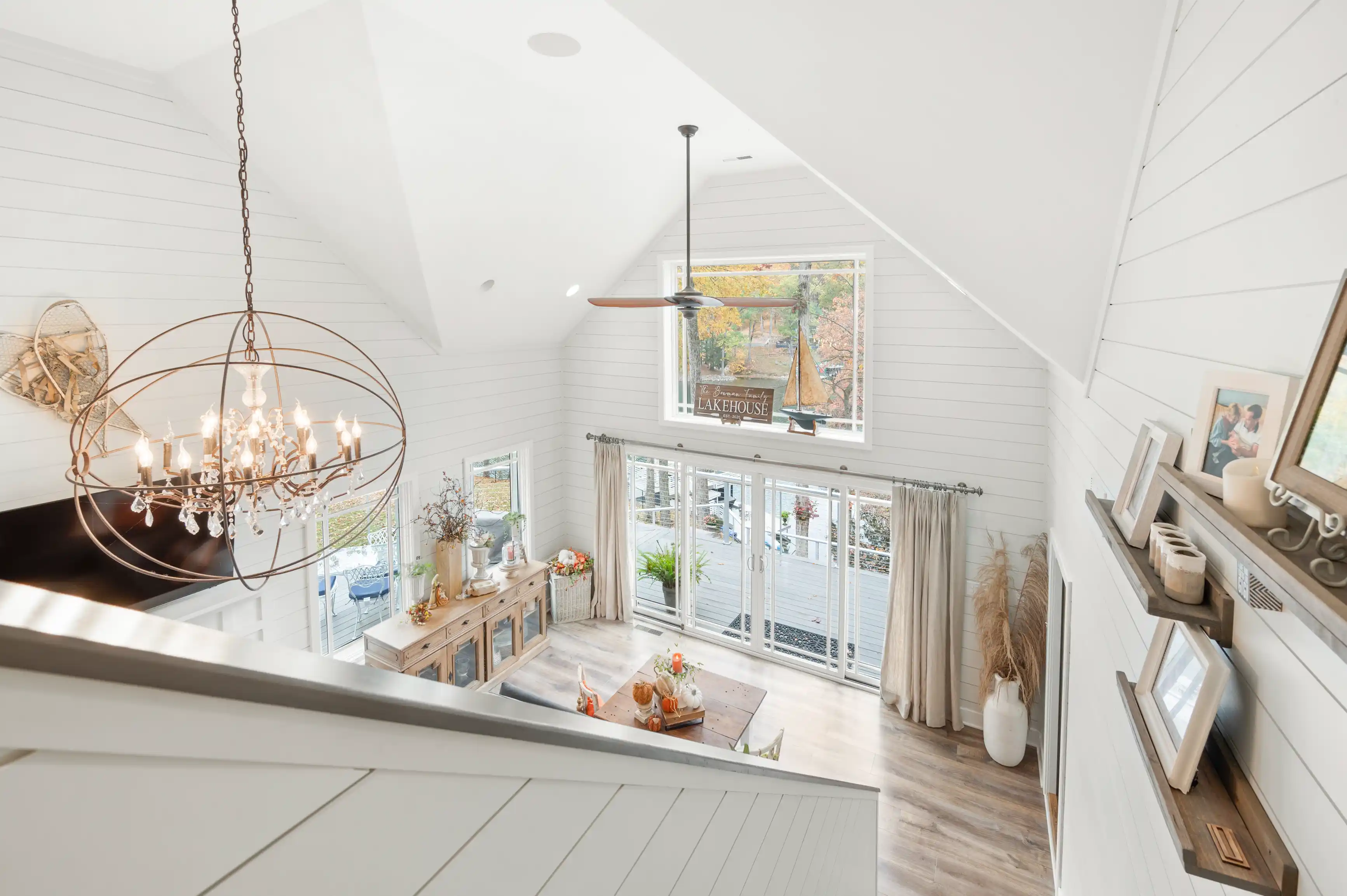 Bright and airy dining room interior in a modern farmhouse style with a wooden table, elegant chandelier, white shiplap walls, and a view of trees through large windows.