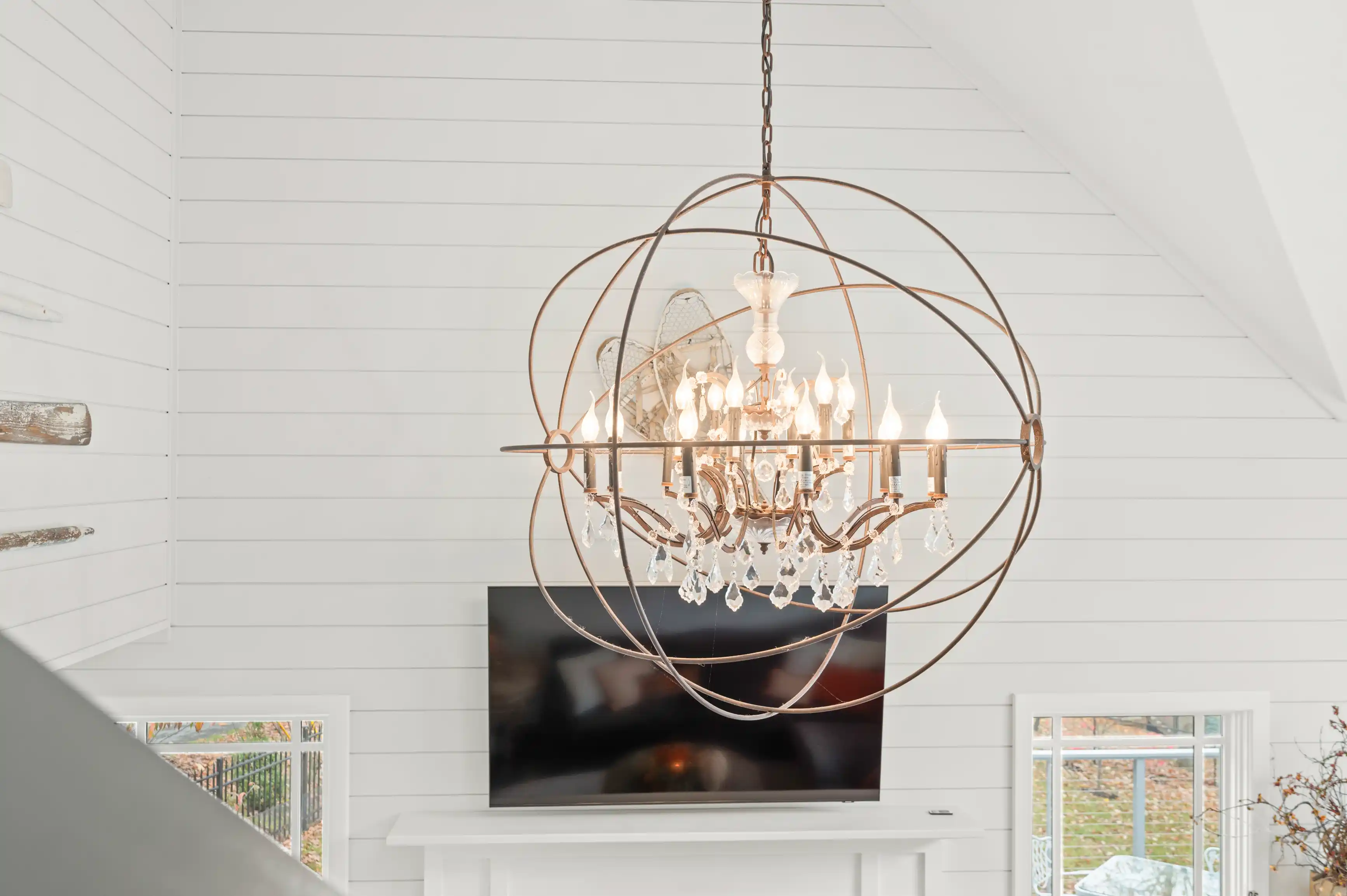 Elegant chandelier with crystal accents hanging in a modern living room with white walls, a vaulted ceiling, and a mounted flat-screen TV visible in the background.