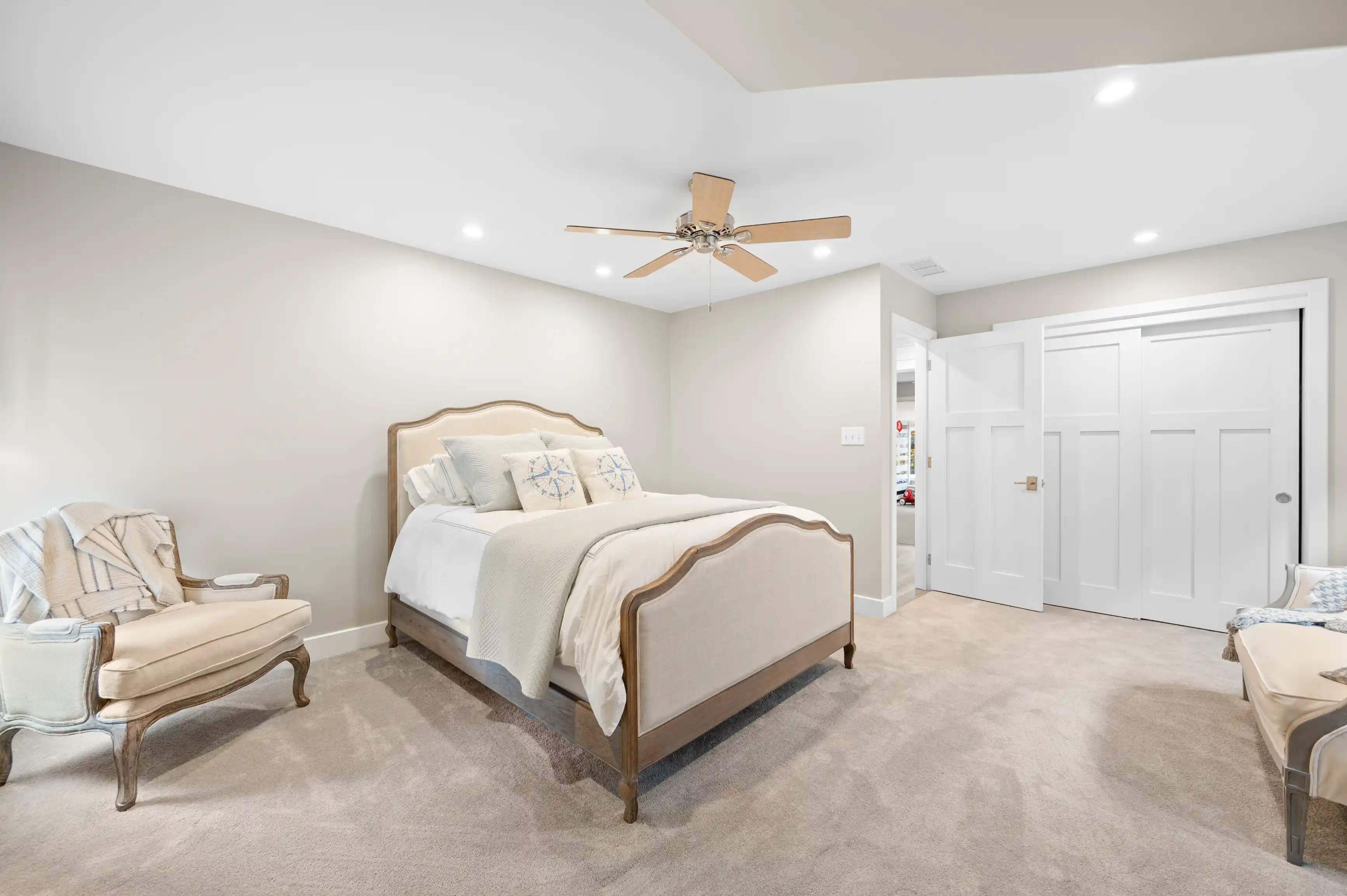 A spacious and well-lit bedroom with a beige upholstered bed, two matching armchairs, a ceiling fan, and a white double door closet.