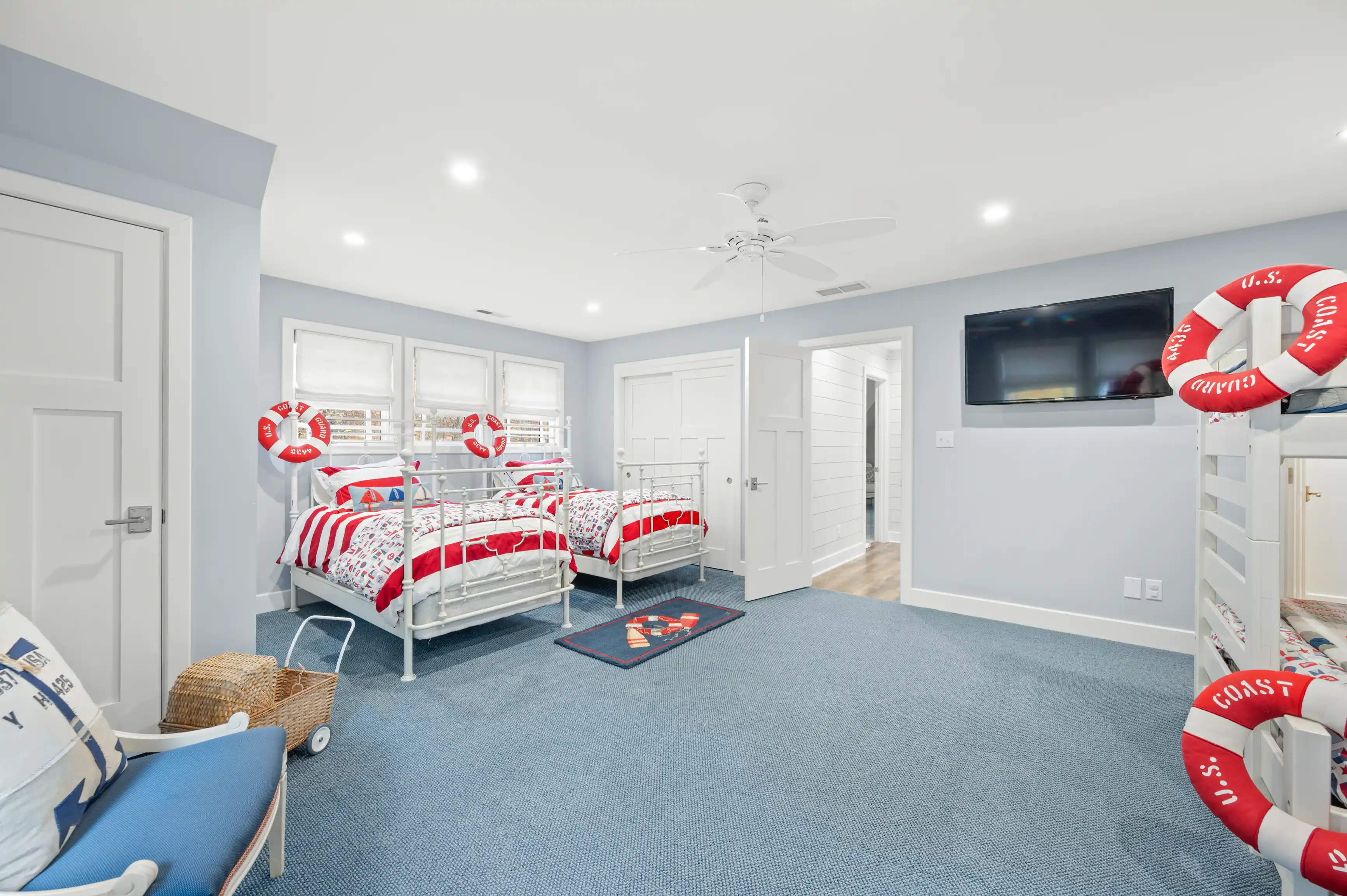 Spacious, nautical-themed bedroom with two twin beds featuring red and white linens, lifebuoy decorations on walls, blue carpet, ceiling fan, flat-screen TV, and a large window letting in natural light.