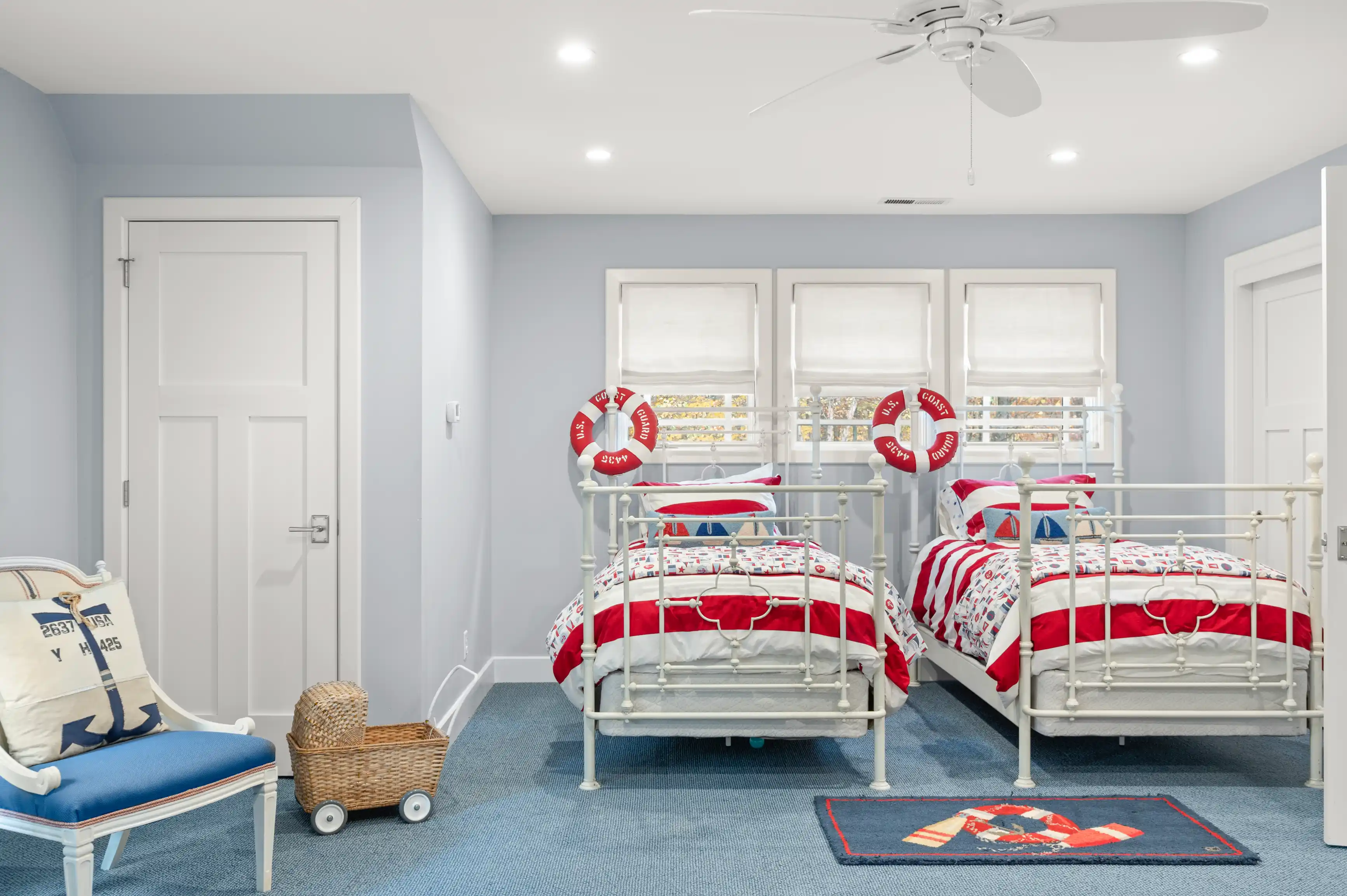 A bright, nautical-themed children's bedroom with two twin beds, life preserver wall decorations, a blue carpet, and a ceiling fan.