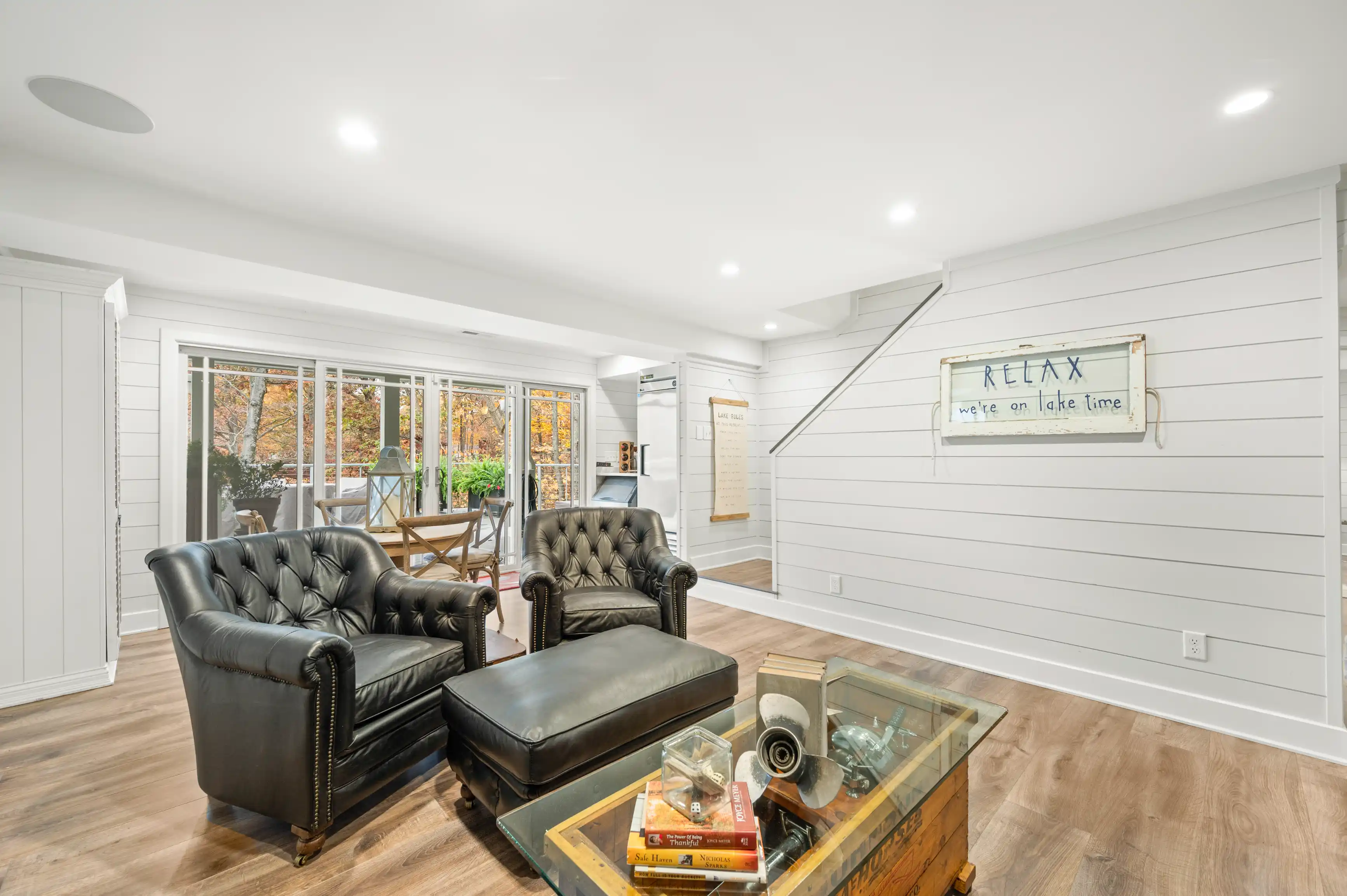 Bright and modern living room with black leather armchairs, a clear coffee table with books, white shiplap walls with a 'RELAX we're on lake time' sign, and sliding glass doors leading to an outdoor deck with autumn foliage visible outside.