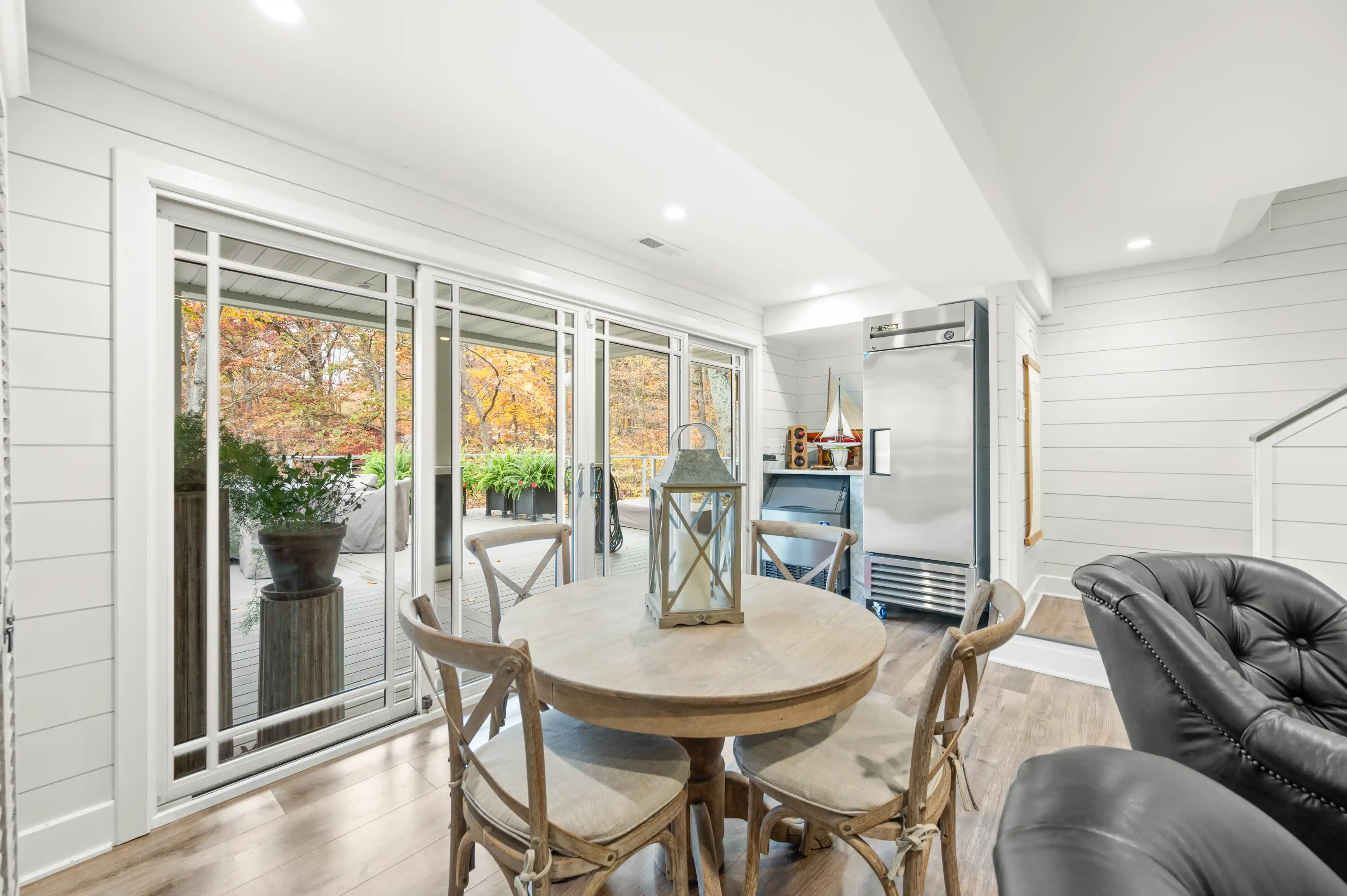 Bright, modern sunroom with sliding glass doors leading to a deck with autumn foliage, featuring a round wooden table, chairs, and a black leather sofa.