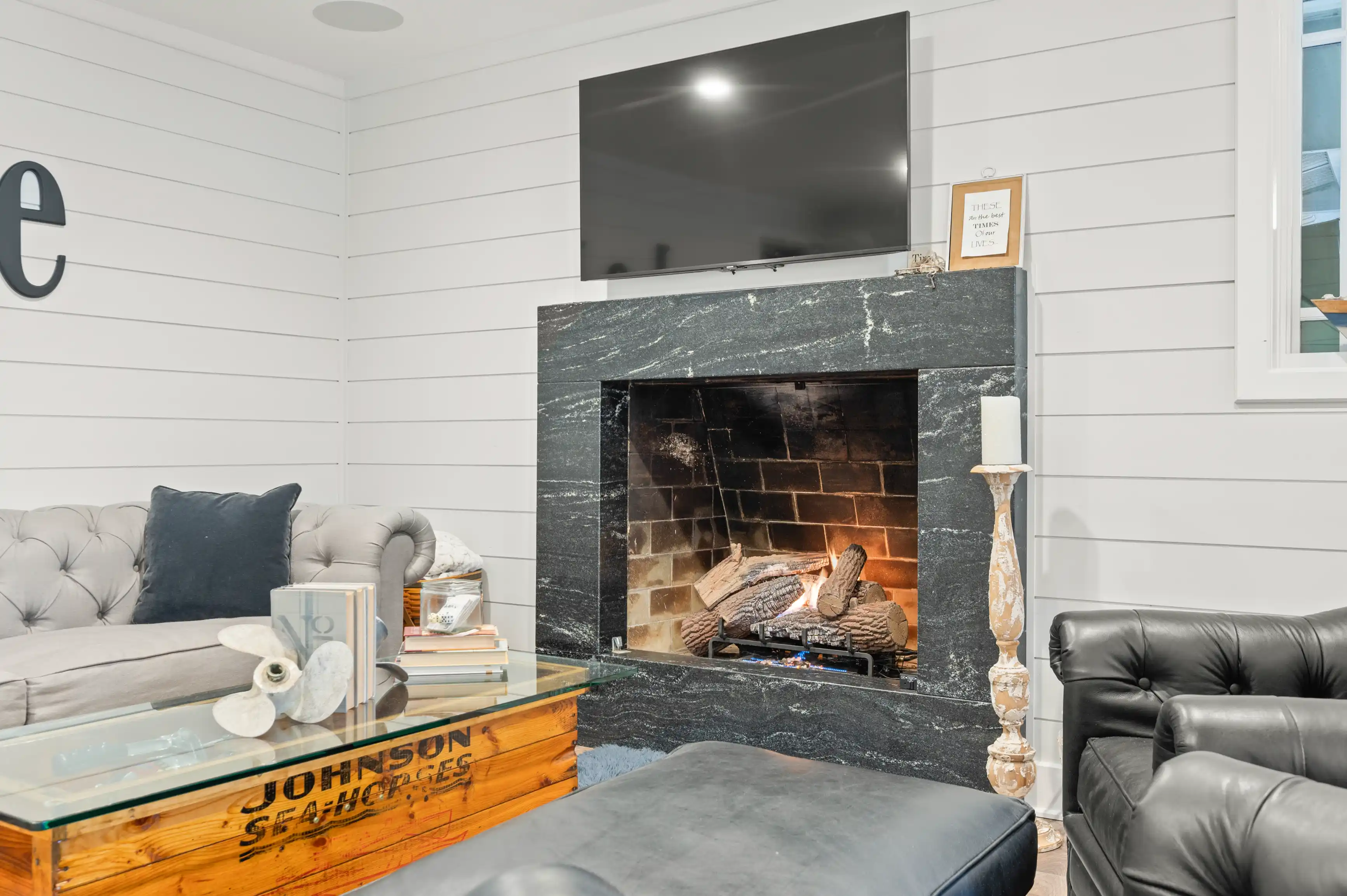 Cozy living room with a lit fireplace, grey tufted sofa, glass coffee table with propeller base, black leather armchair, and shiplap walls with mounted TV above the fireplace.