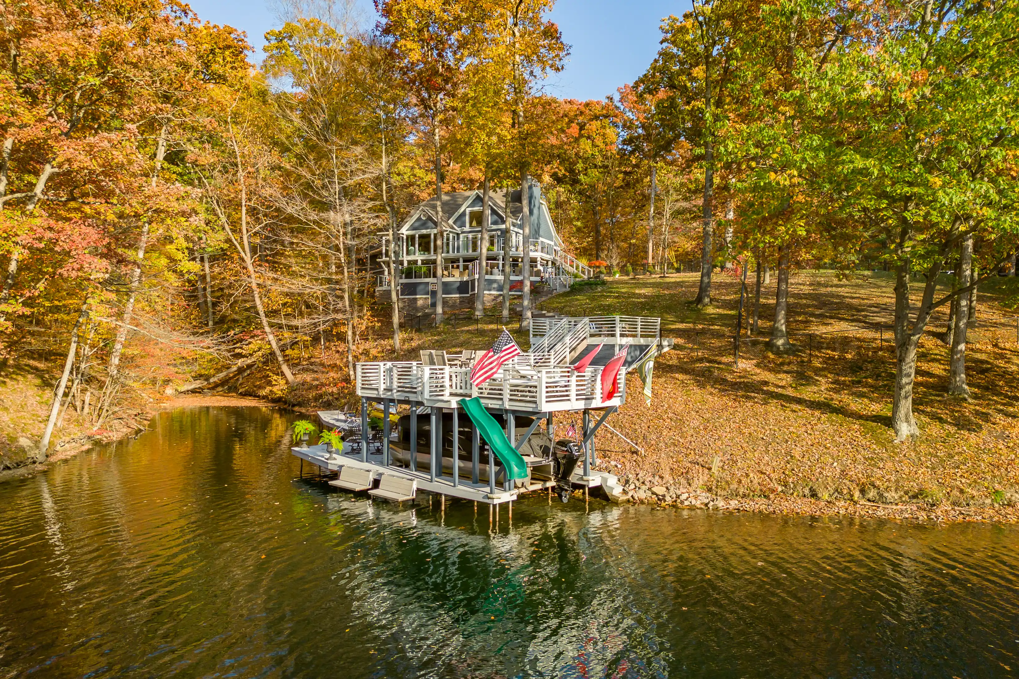 A lakeside house surrounded by autumn-colored trees with a dock featuring lounge chairs, American flags, and a green slide.