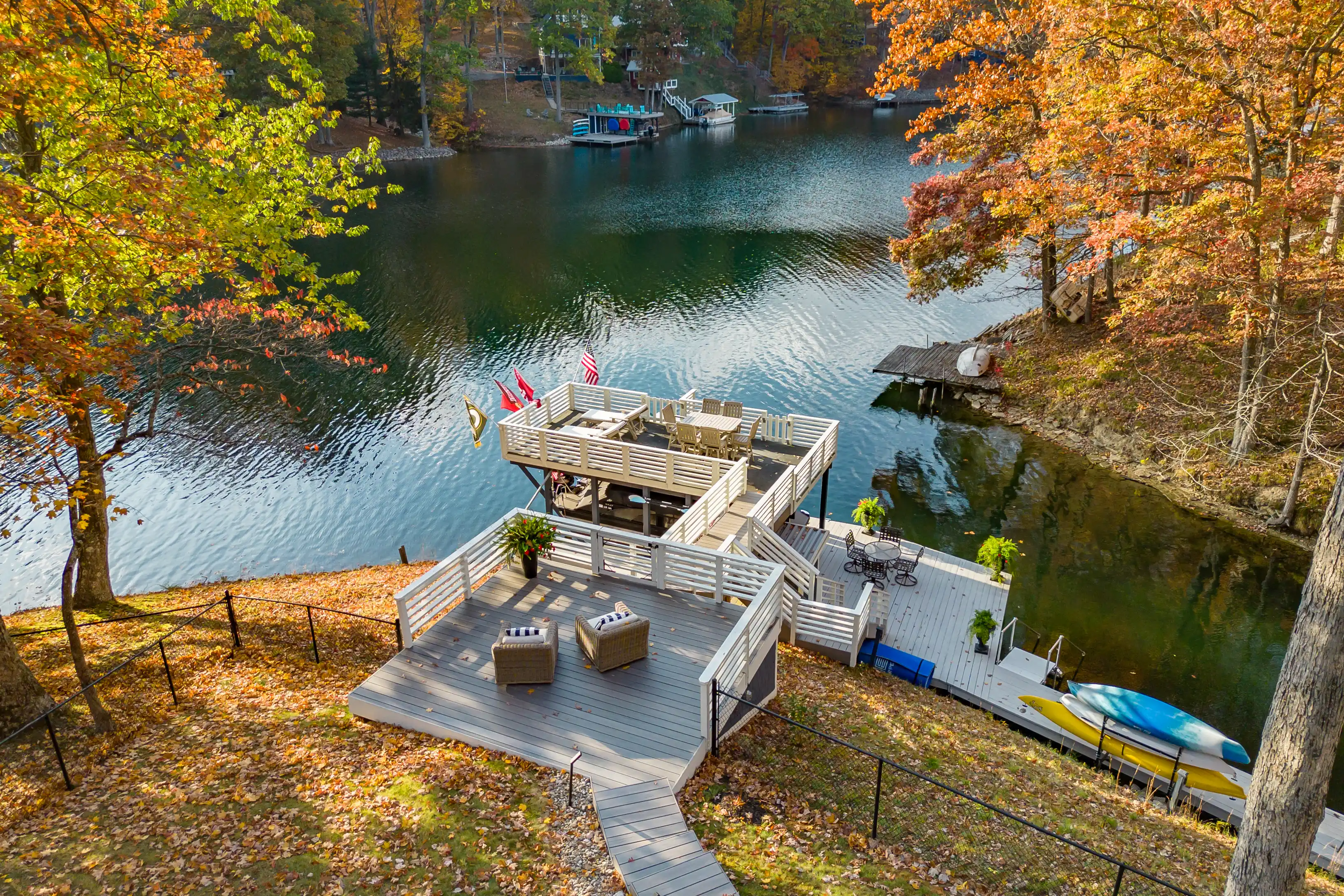 Aerial view of a lakeside wooden deck with outdoor furniture surrounded by trees with autumn foliage, a jet ski, and a boat docked nearby.