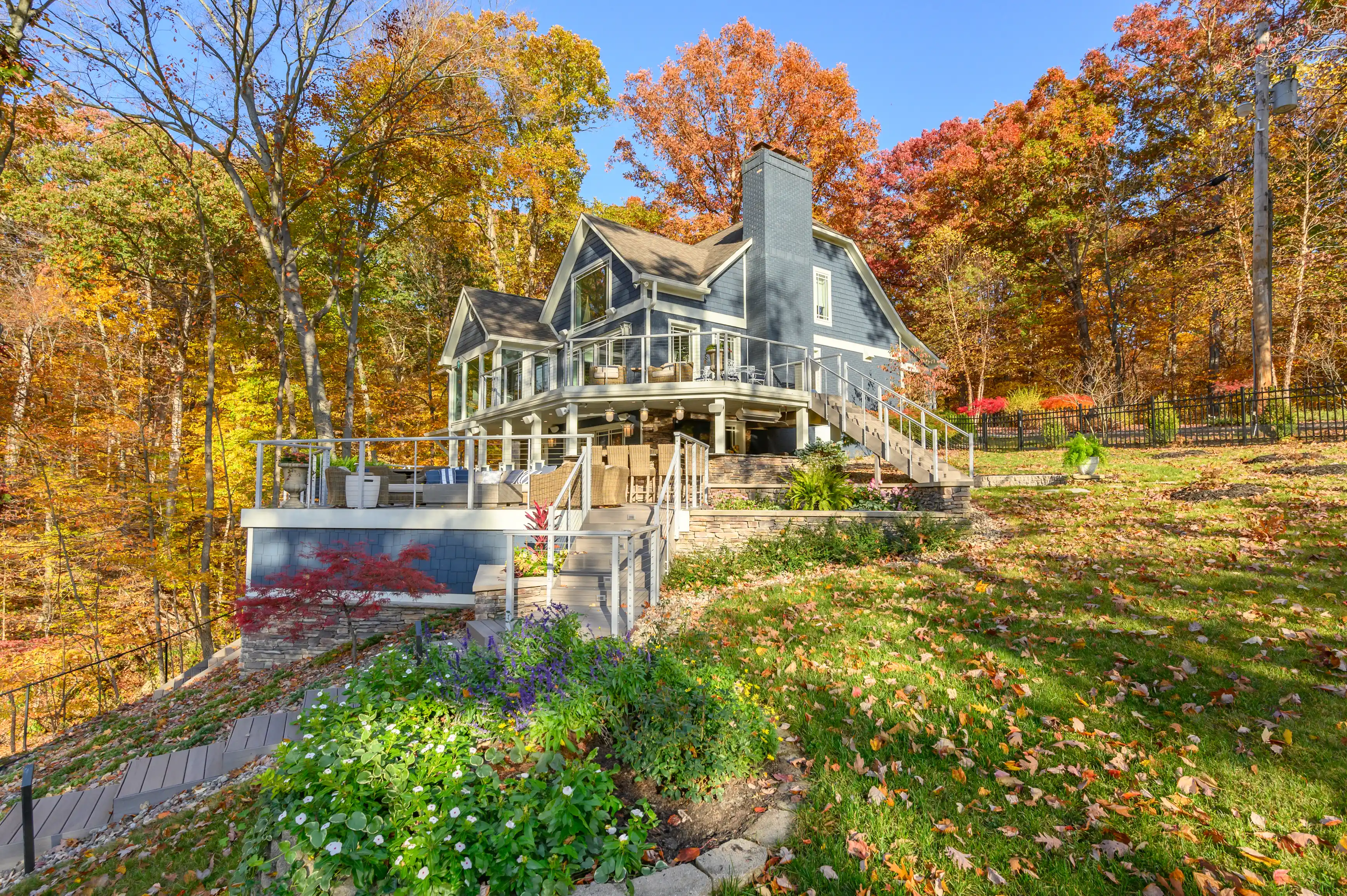 A large two-story house with a wraparound porch surrounded by trees with autumn foliage and a landscaped garden in the foreground.
