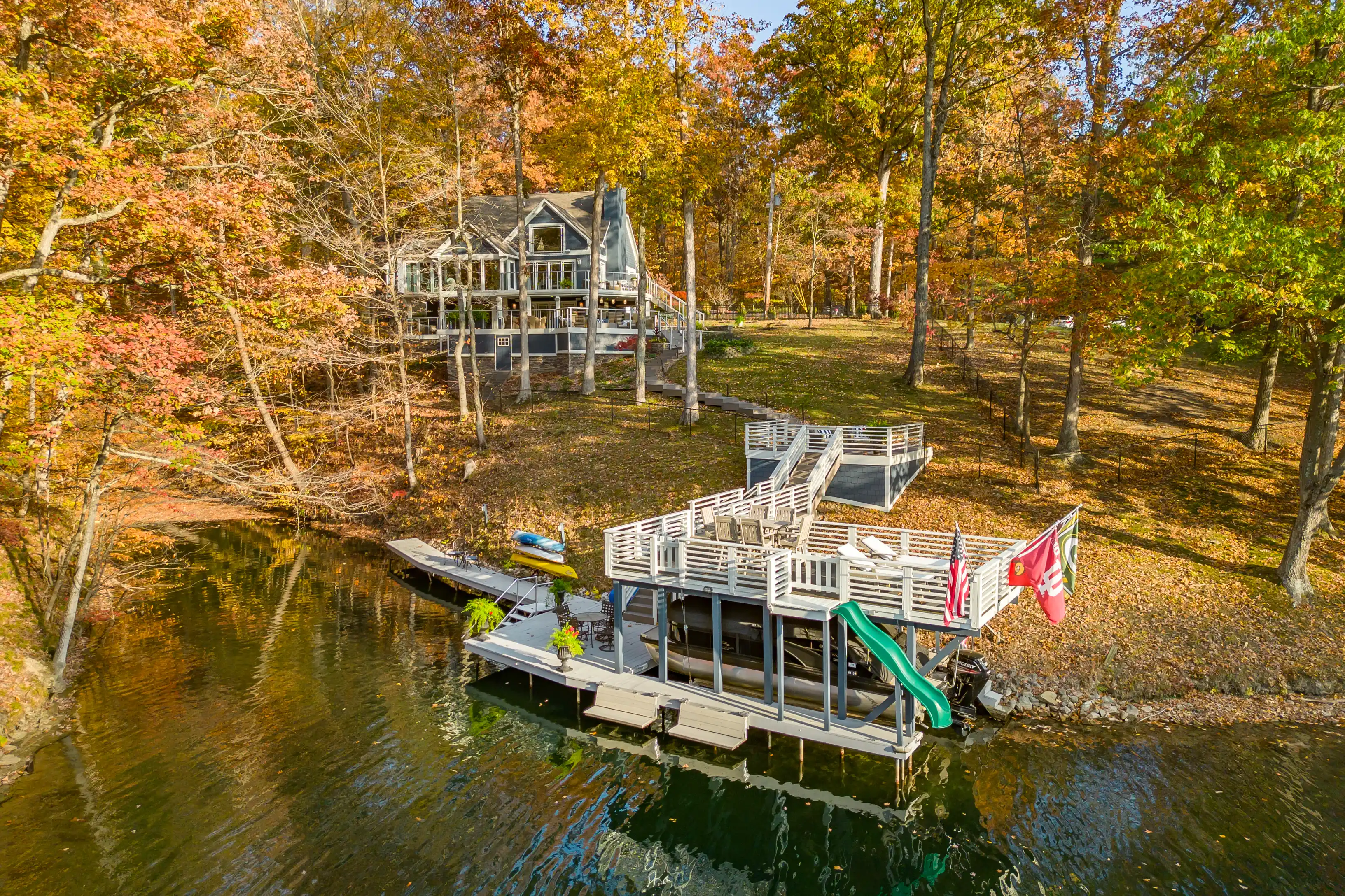 Aerial view of a lake house with a large wooden pier and a double-decker dock, surrounded by trees with autumn foliage.