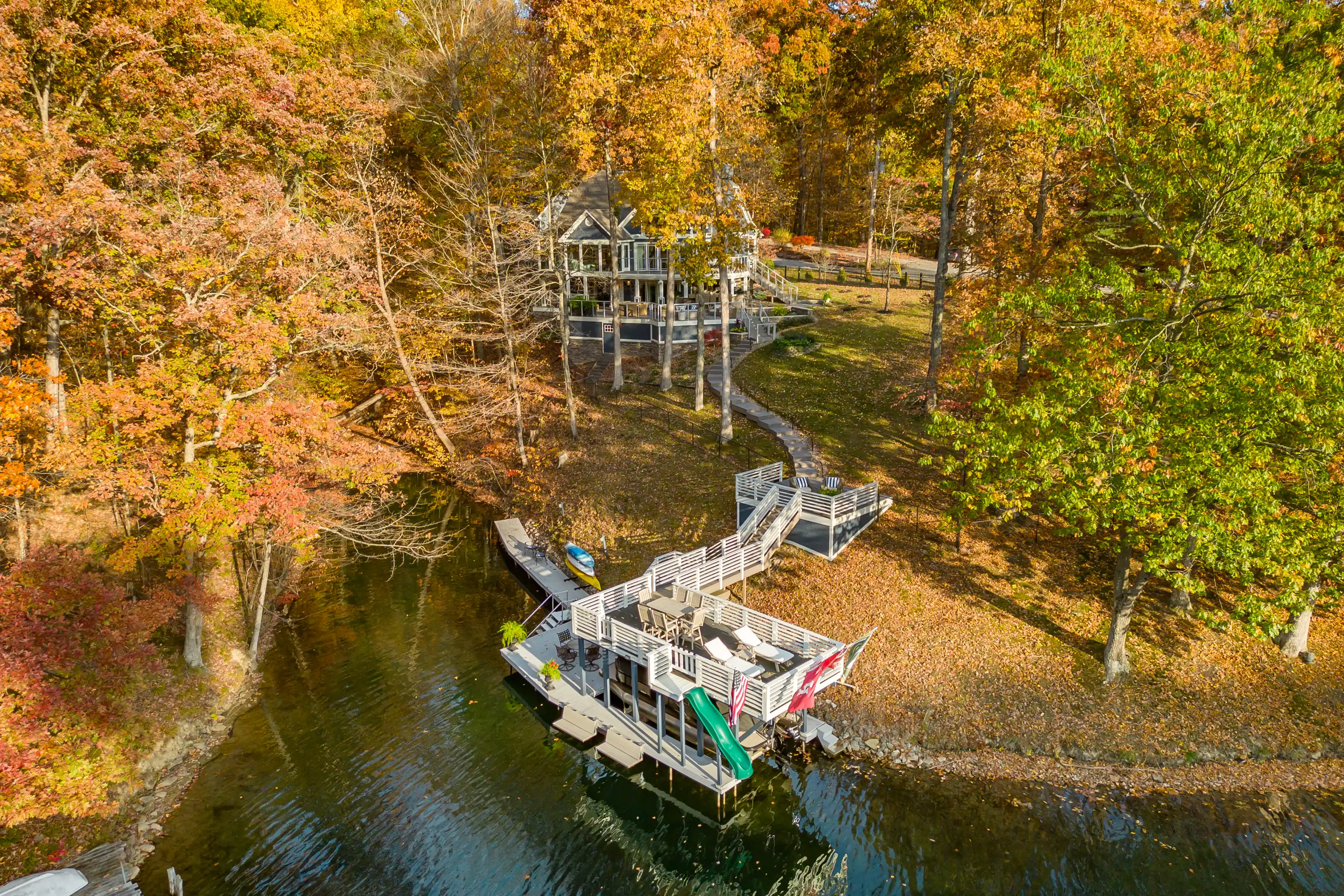 Aerial view of a lakeside house with a dock during autumn, surrounded by trees with colorful foliage.