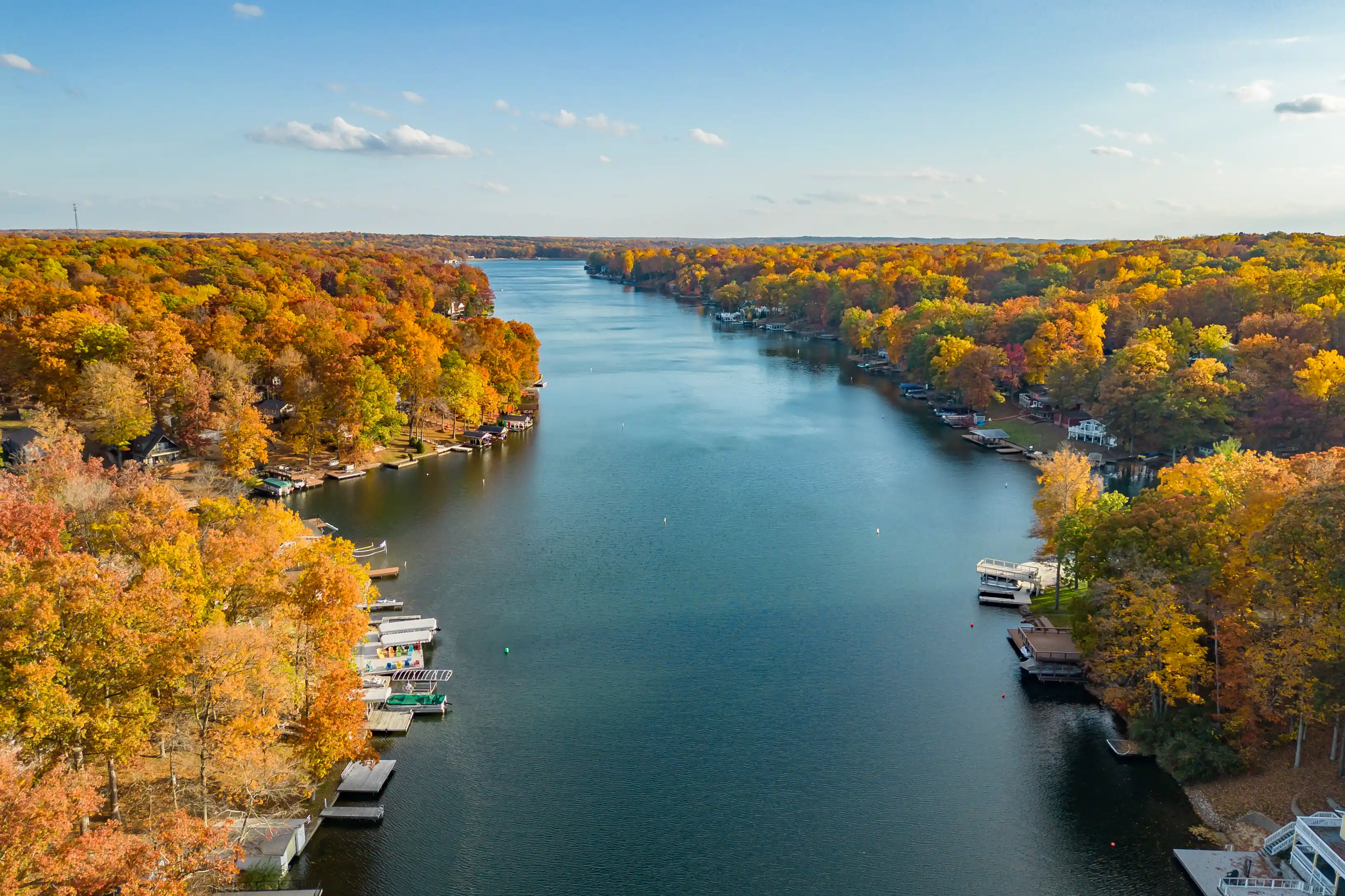Aerial view of a river surrounded by trees with autumn foliage, with docks and boats along the banks.