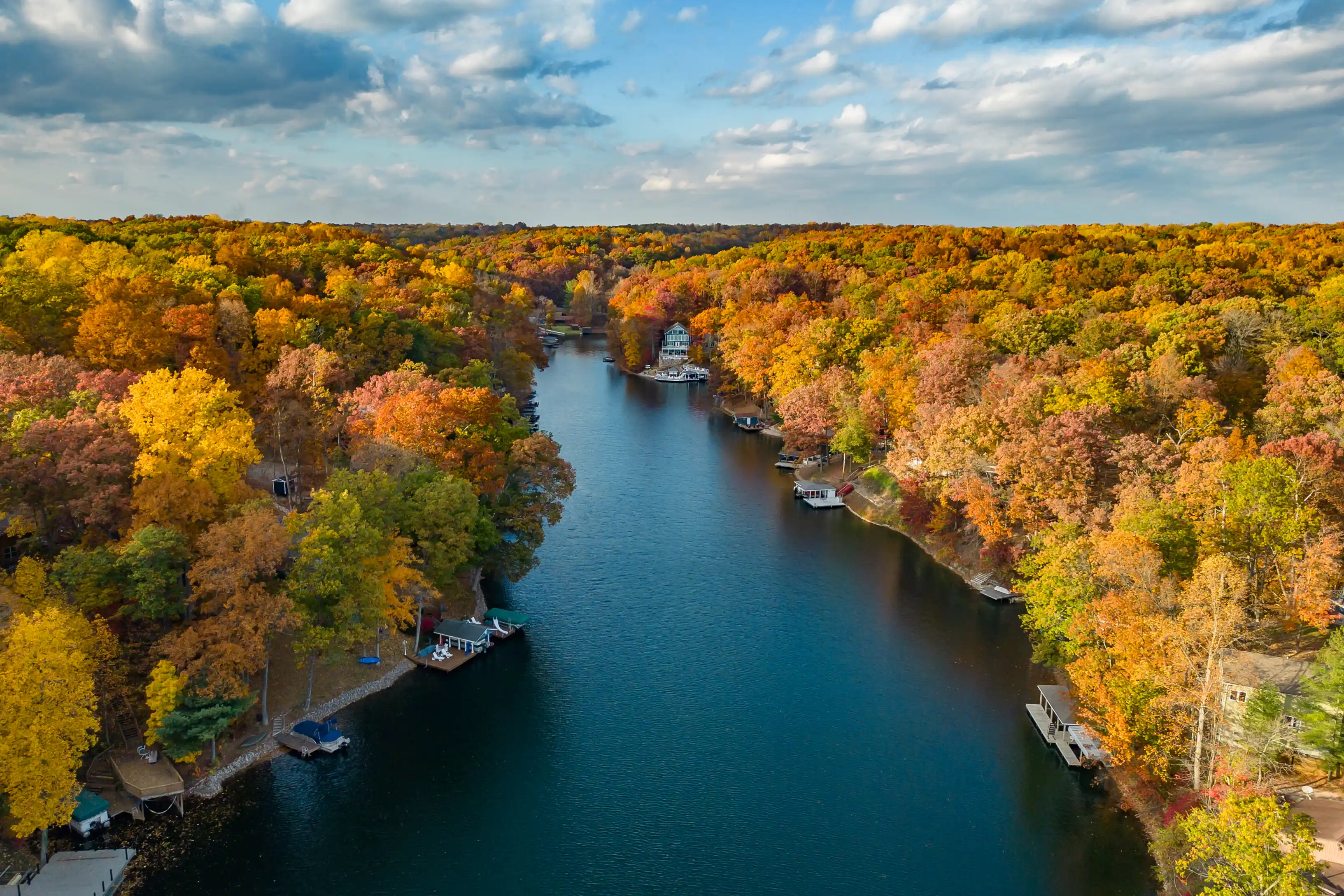 Aerial view of a river flanked by trees with vibrant autumn foliage and docks with boats near the banks.
