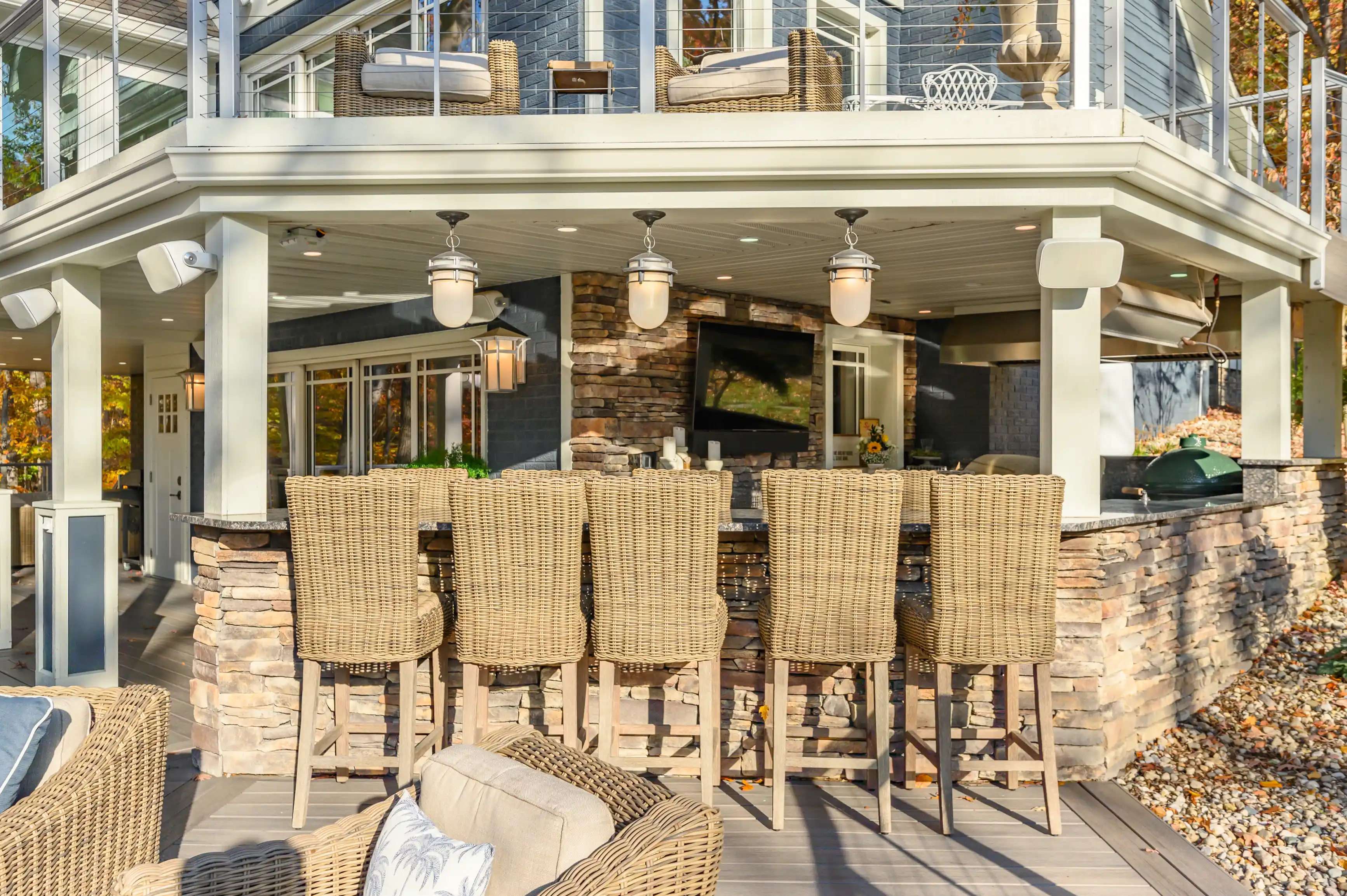 Elegant outdoor patio area with wicker bar chairs, a stone fireplace, and a built-in barbecue grill under a covered deck.