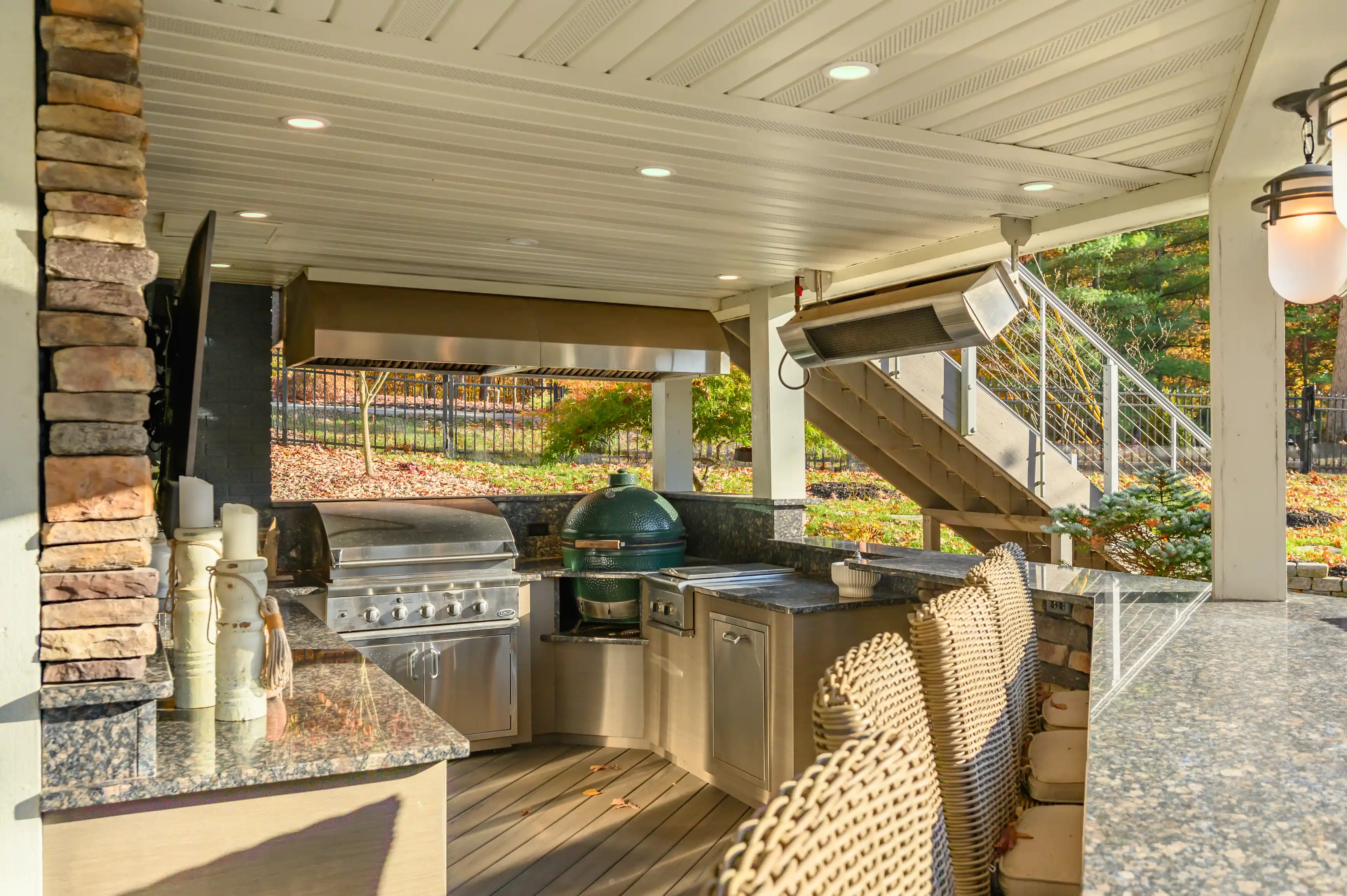 Outdoor patio with modern grilling area, including a built-in barbecue and smoker, under a covered deck with wicker furniture, overlooking an autumn garden.