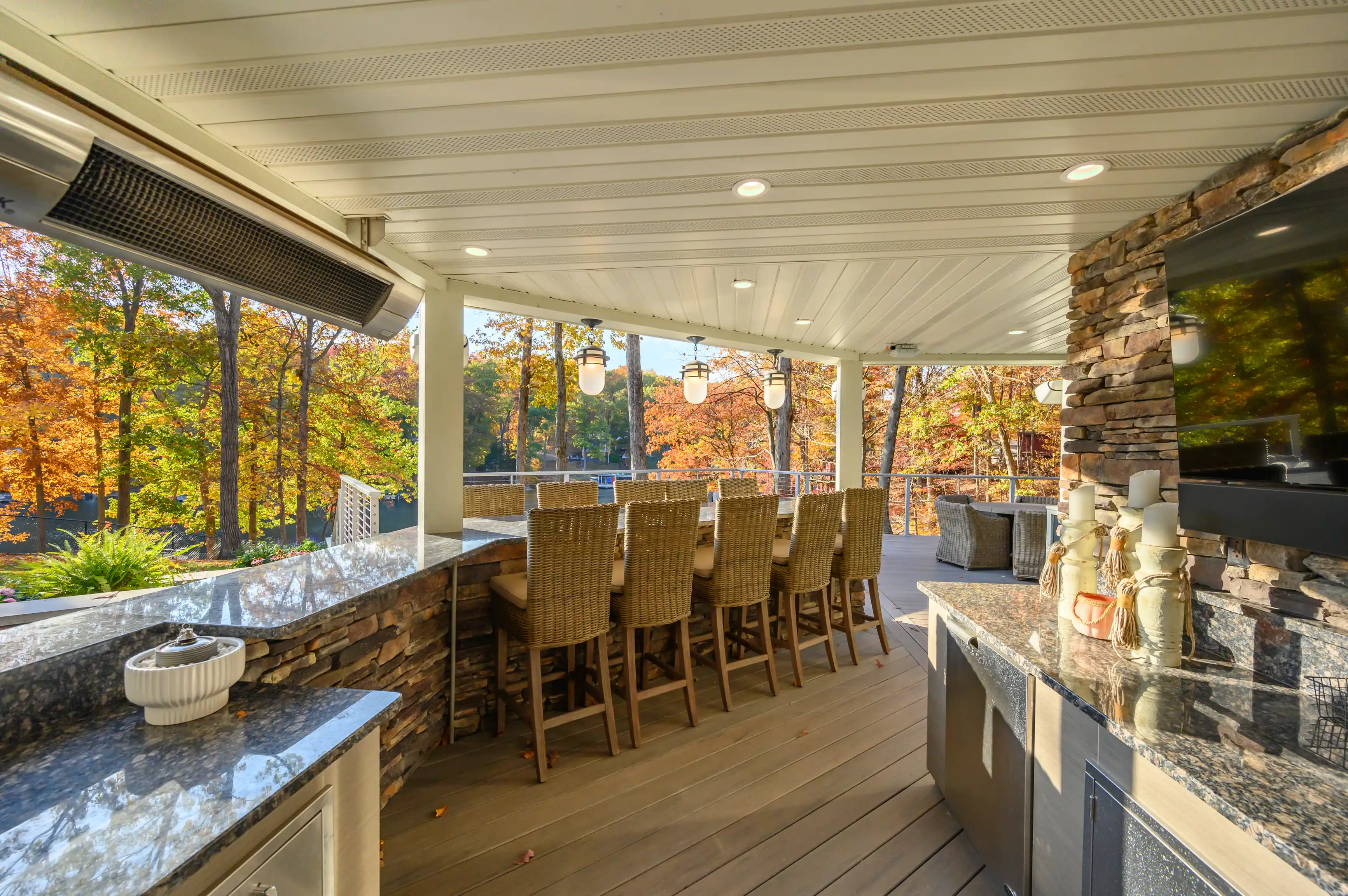 Elegant outdoor covered patio area with wicker bar stools, a stone fireplace, and a view of autumn trees.