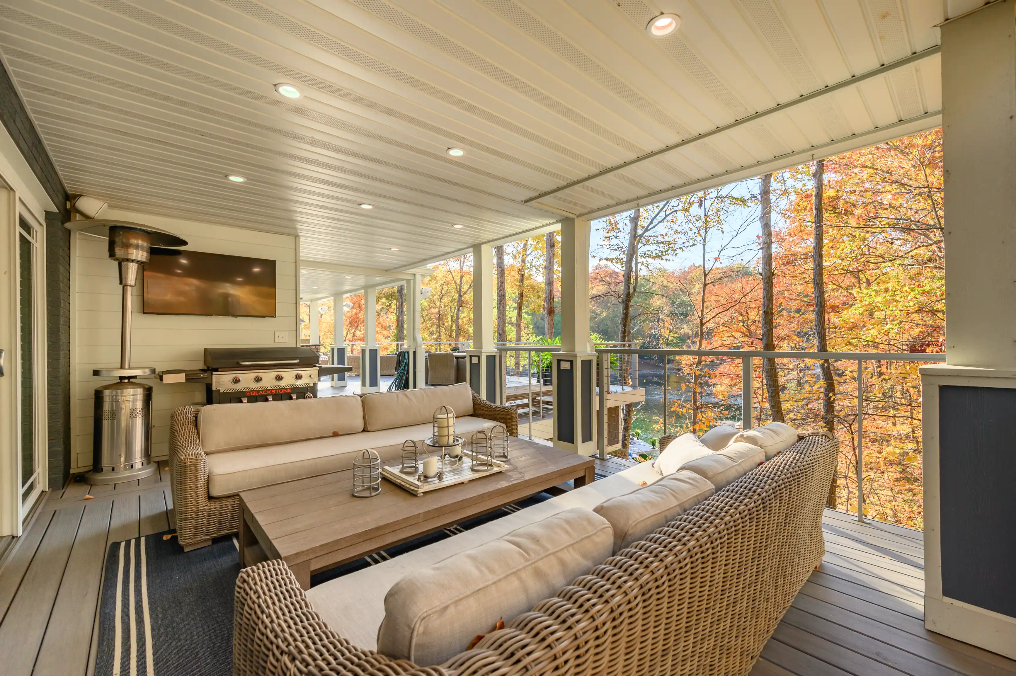 Spacious covered outdoor deck with comfortable seating, a grill, and a picturesque view of autumn trees.