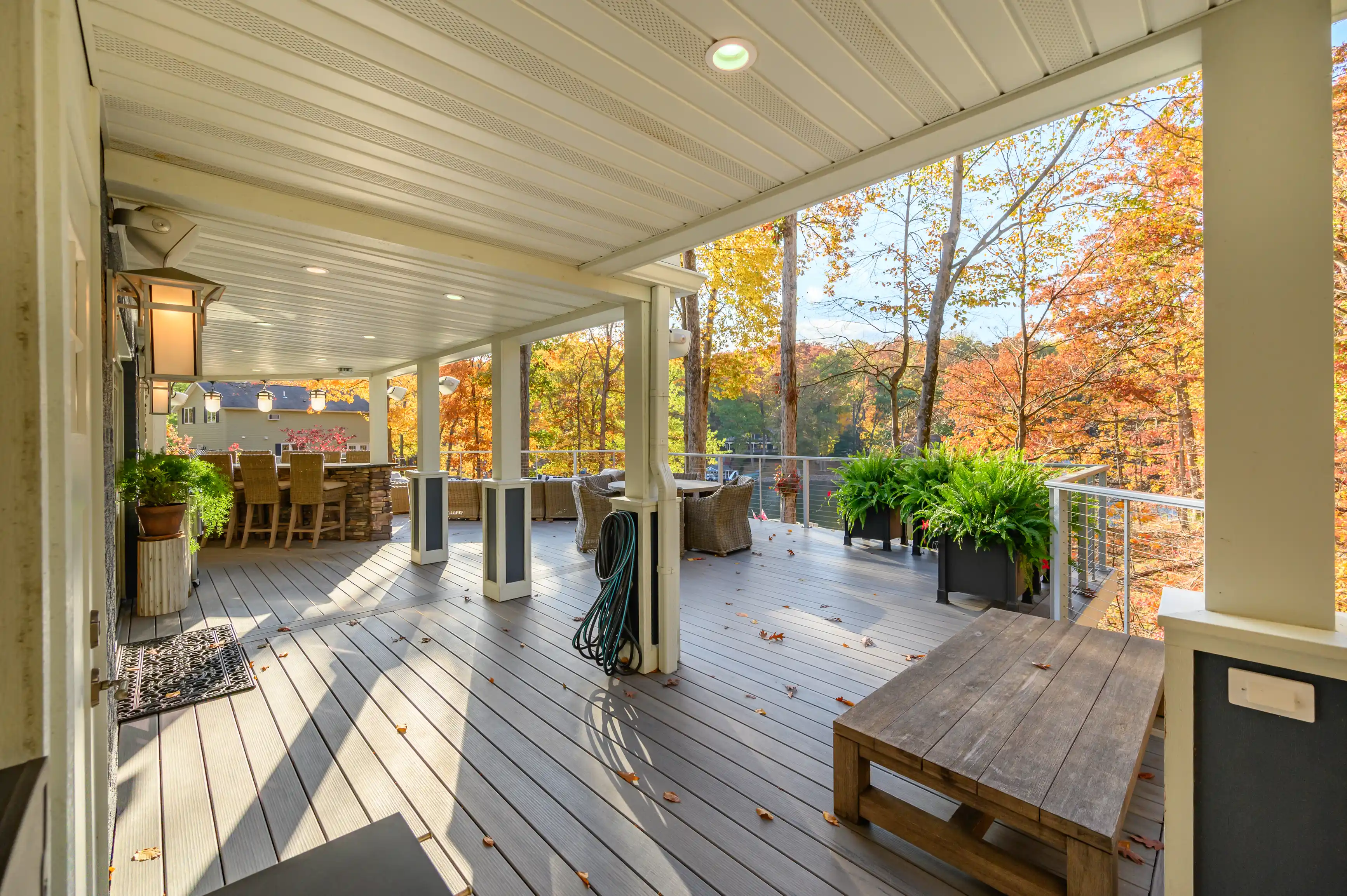 Spacious deck with outdoor seating surrounded by autumn foliage, featuring potted plants and a wooden bench under a covered area with a beadboard ceiling.