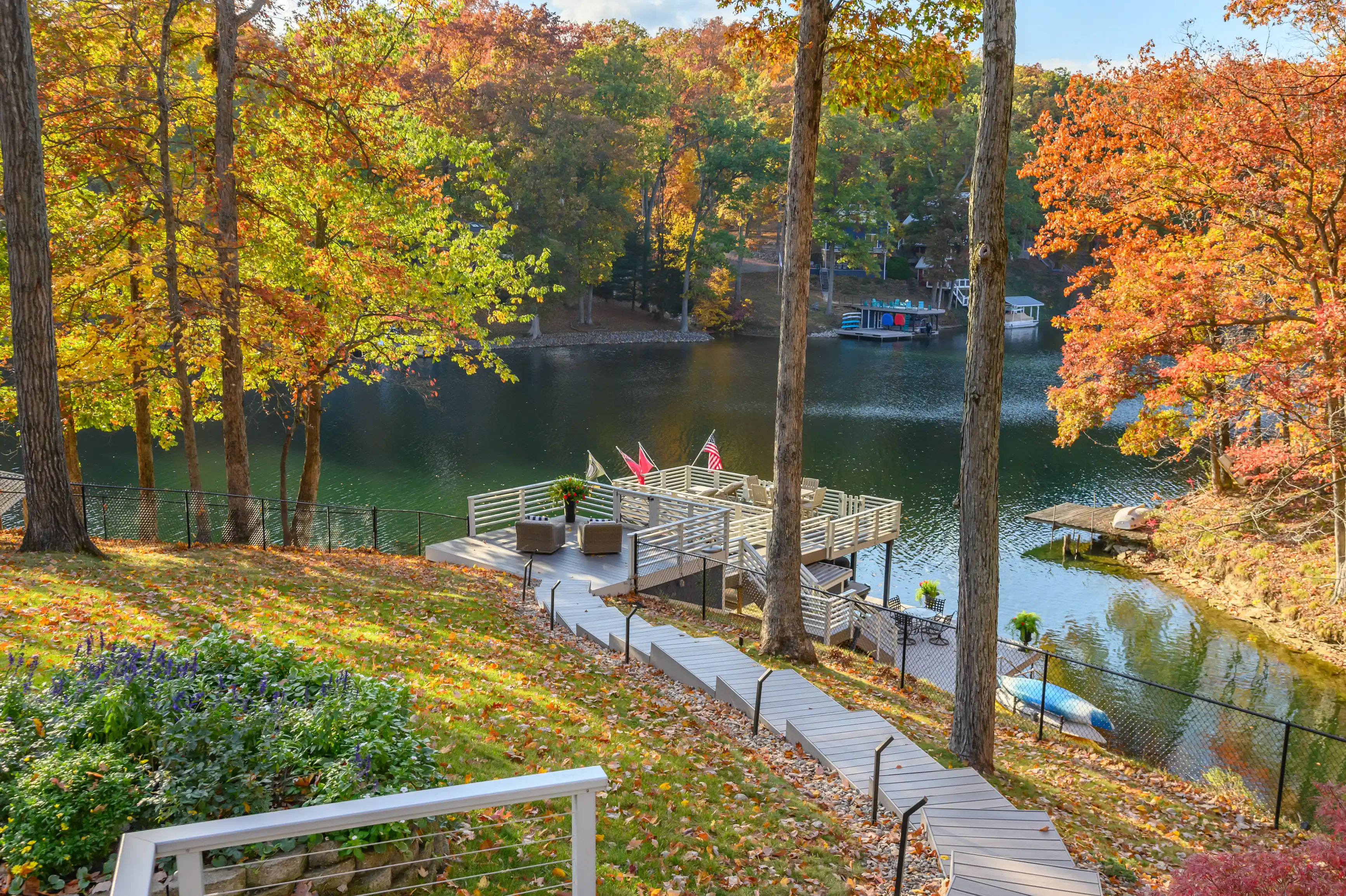 Autumnal view of a backyard leading down to a lake with a dock, outdoor furniture, surrounded by trees with colorful fall foliage.