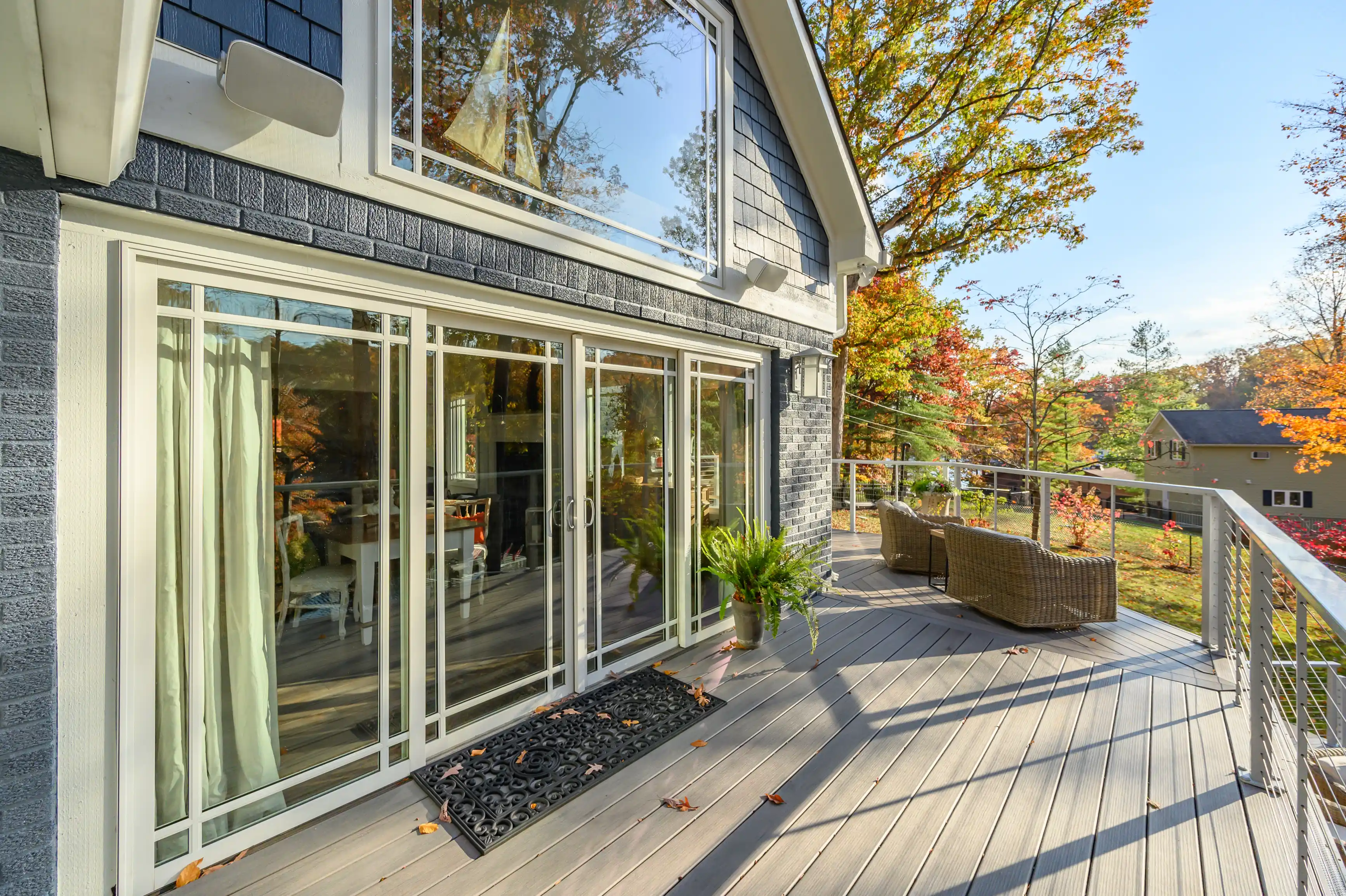 Modern house exterior with a spacious deck featuring sliding glass doors, wicker chairs, and autumn foliage in the background.