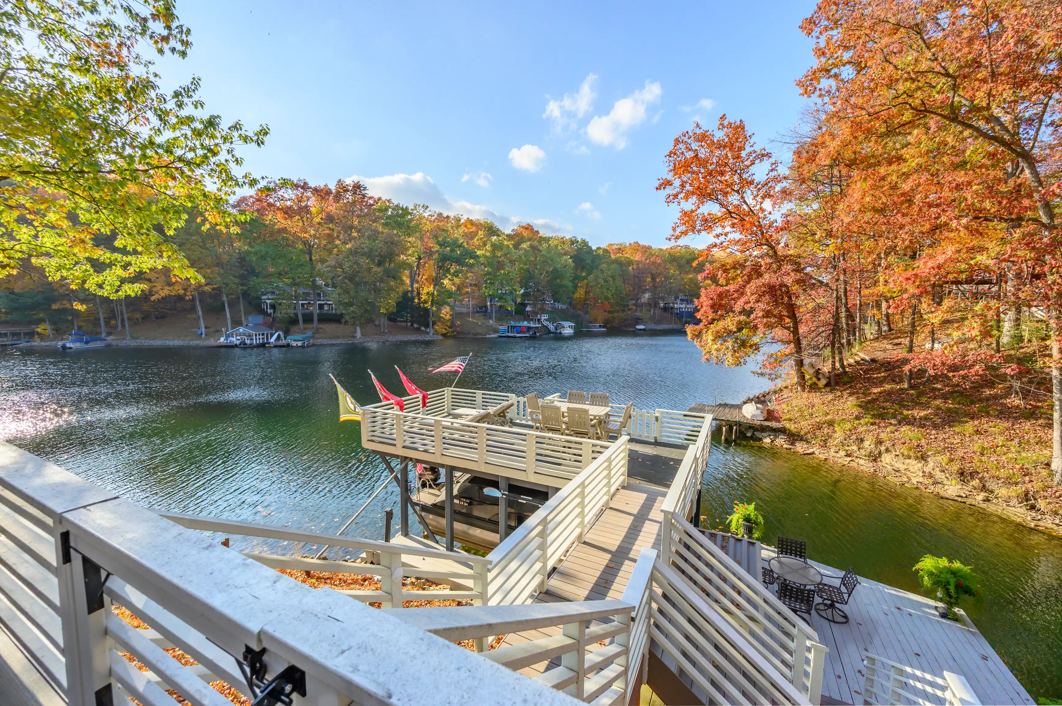 A scenic view from a deck overlooking a lake surrounded by autumn-colored trees, with a boat dock and boats in the distance.