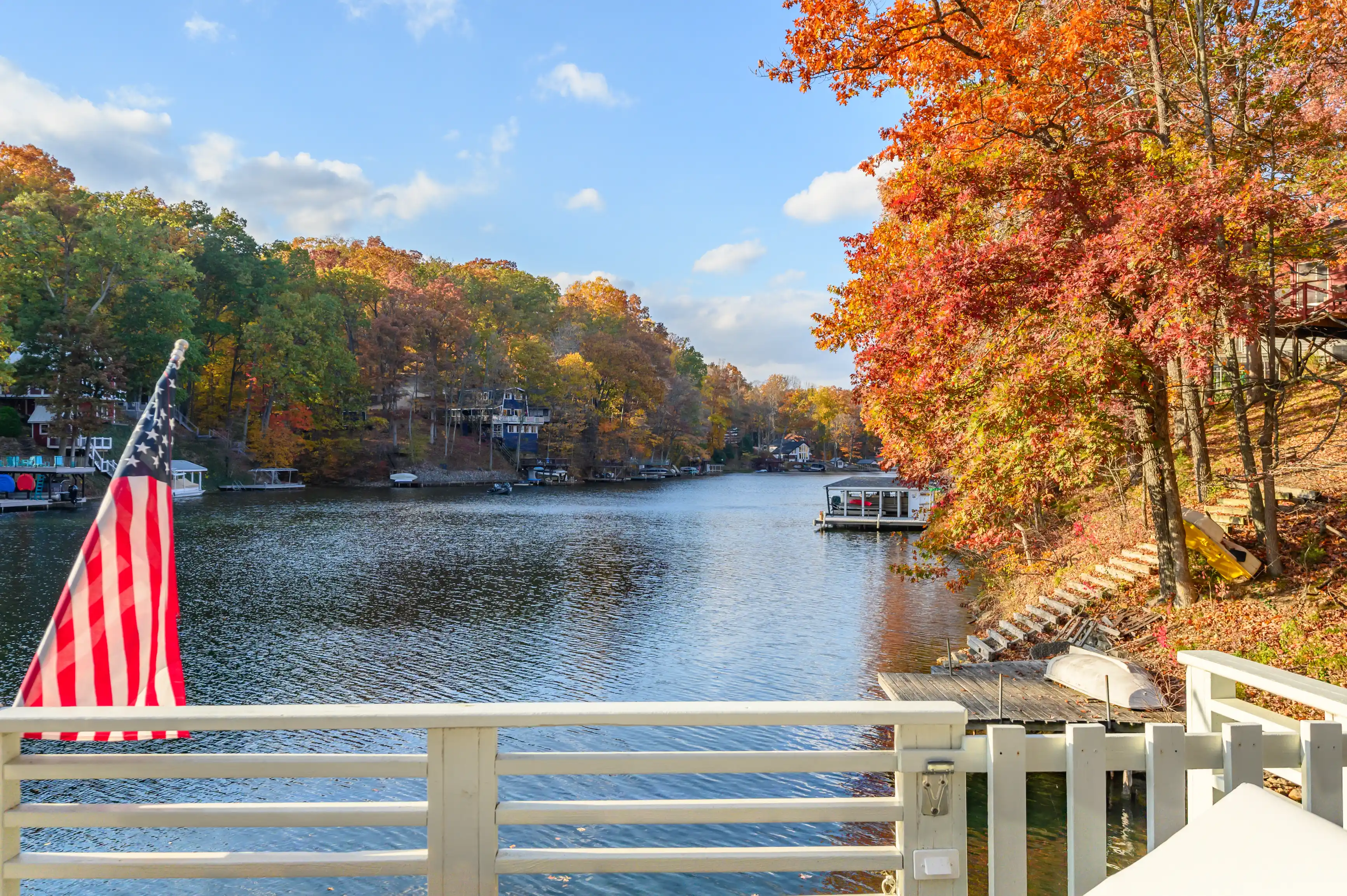 A serene autumn lakeside view with colorful trees, an American flag in the foreground, and houses dotted along the shore.