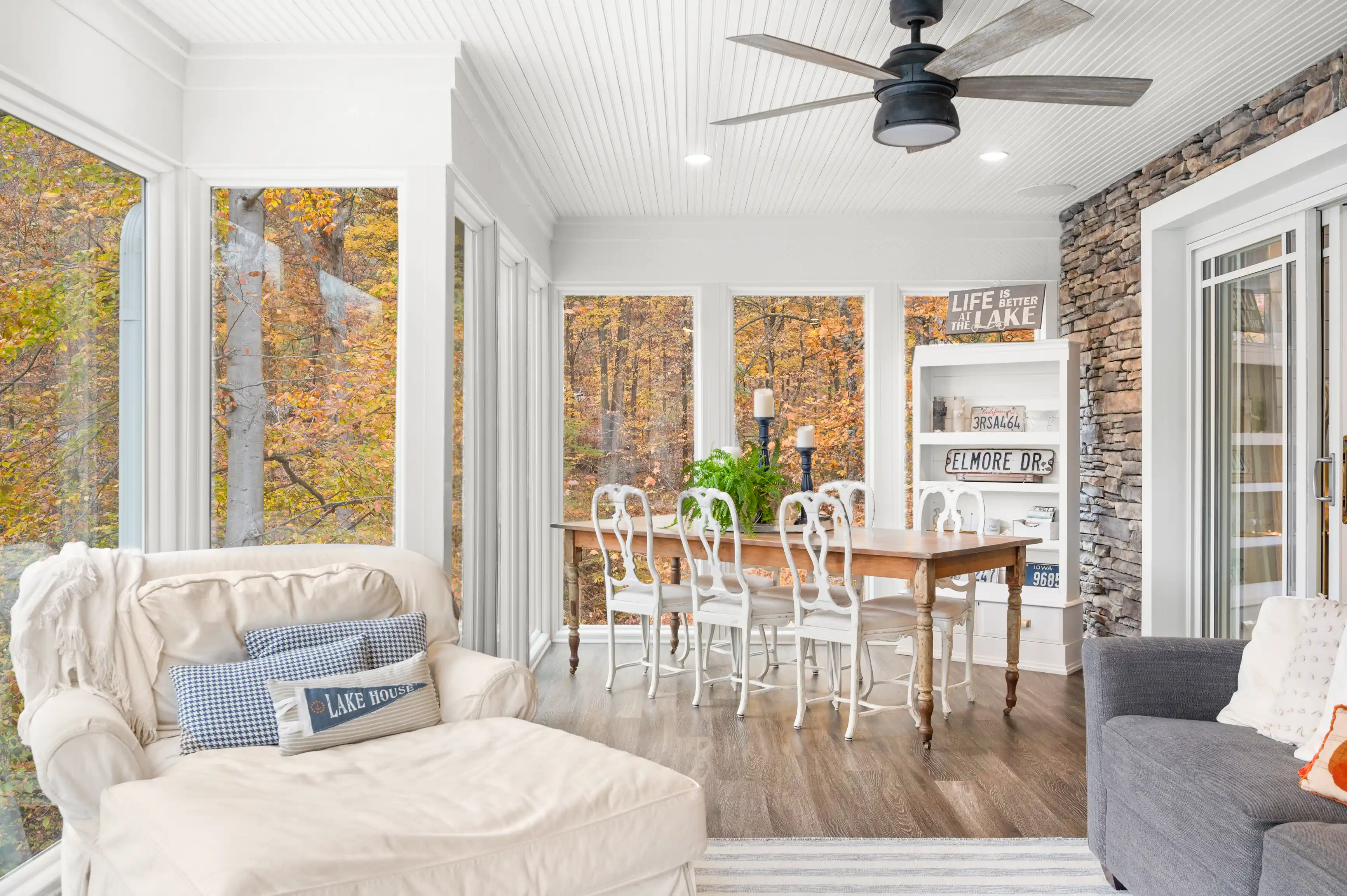 Cozy lakeside home interior with large windows showing autumn trees, a dining area with a wooden table and white chairs, and a living space with comfortable sofas, decorative cushions, and a ceiling fan.