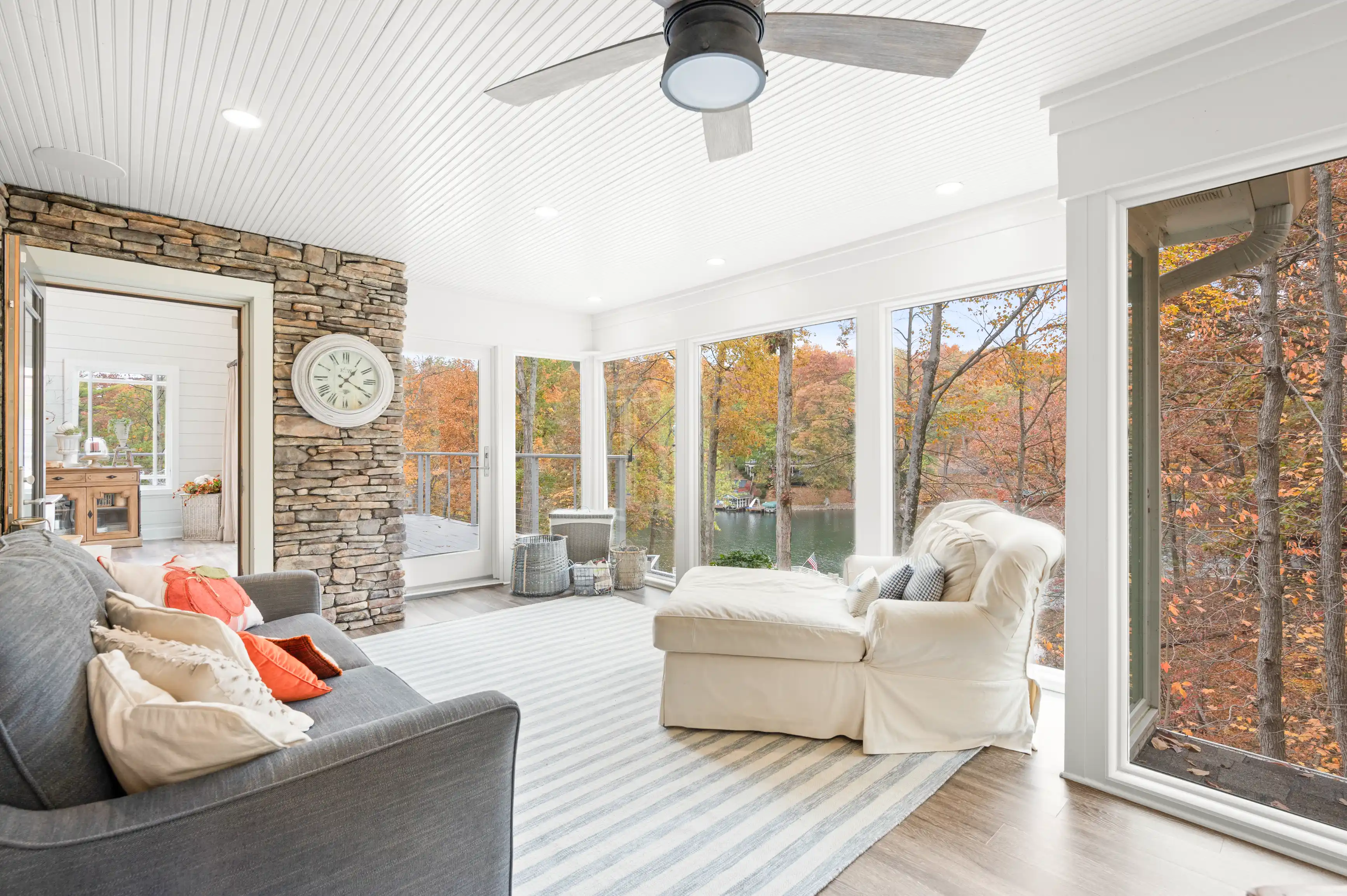 Bright and airy sunroom with large windows showing fall foliage, stone fireplace, comfortable seating, and ceiling fan.