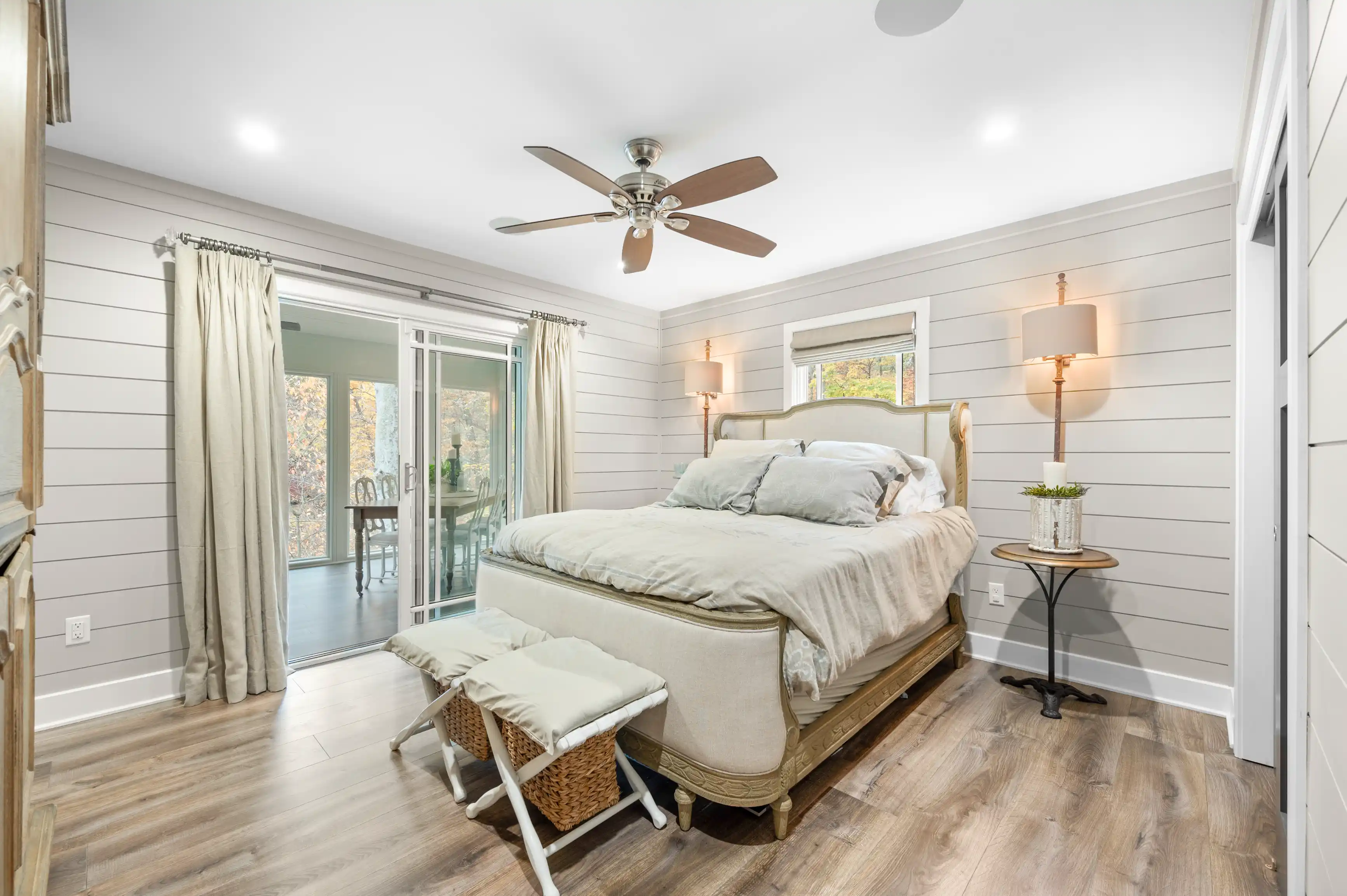Bright and modern bedroom with a queen-sized bed, two bedside lamps, a ceiling fan, a folding chair with a cushion, sliding glass doors leading to an outdoor area, and shiplap walls.