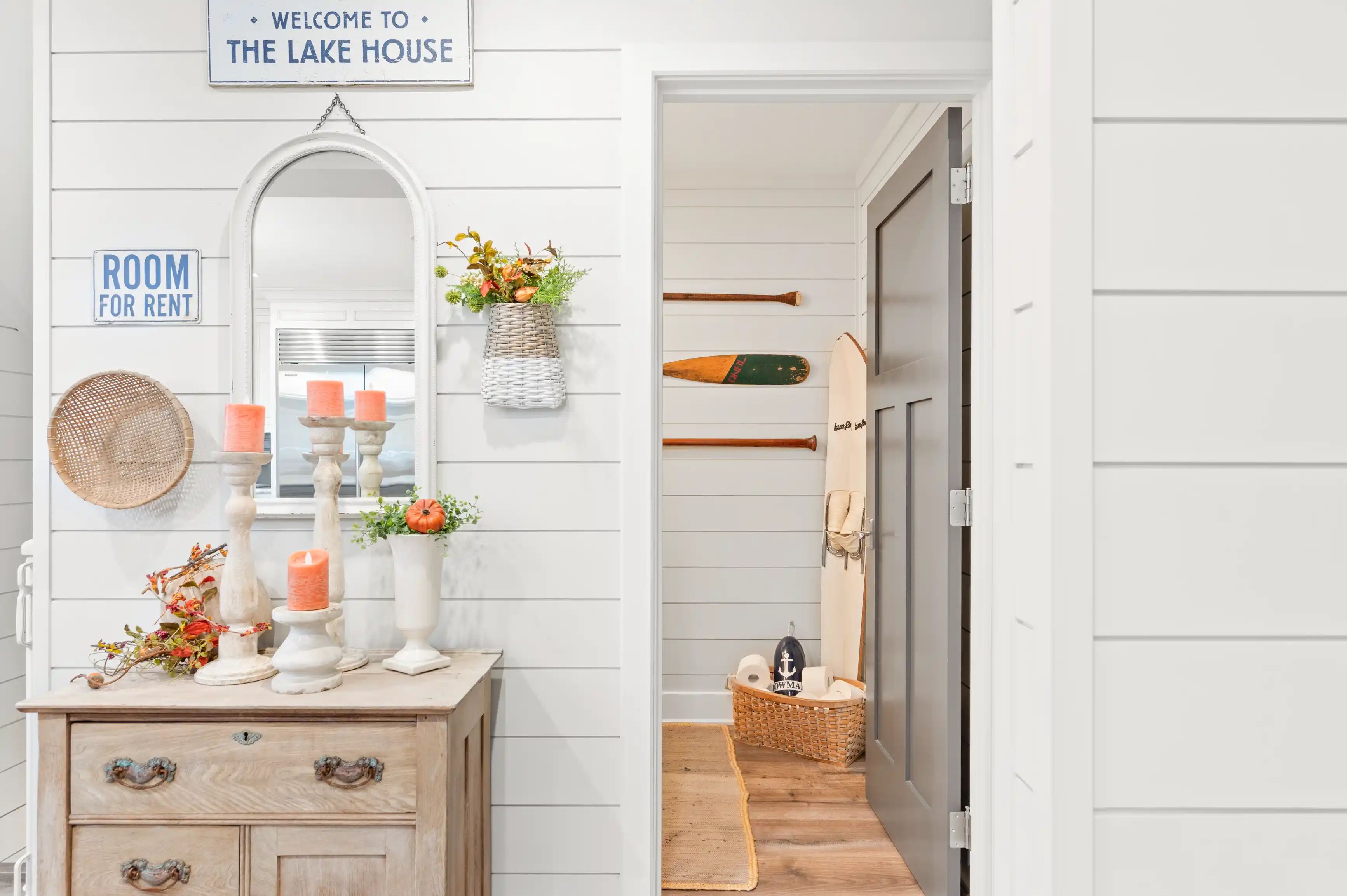 Cozy lake house entryway with a wooden dresser, mirror, decorative candles, wall-mounted oars, and a sign reading "Welcome to The Lake House".